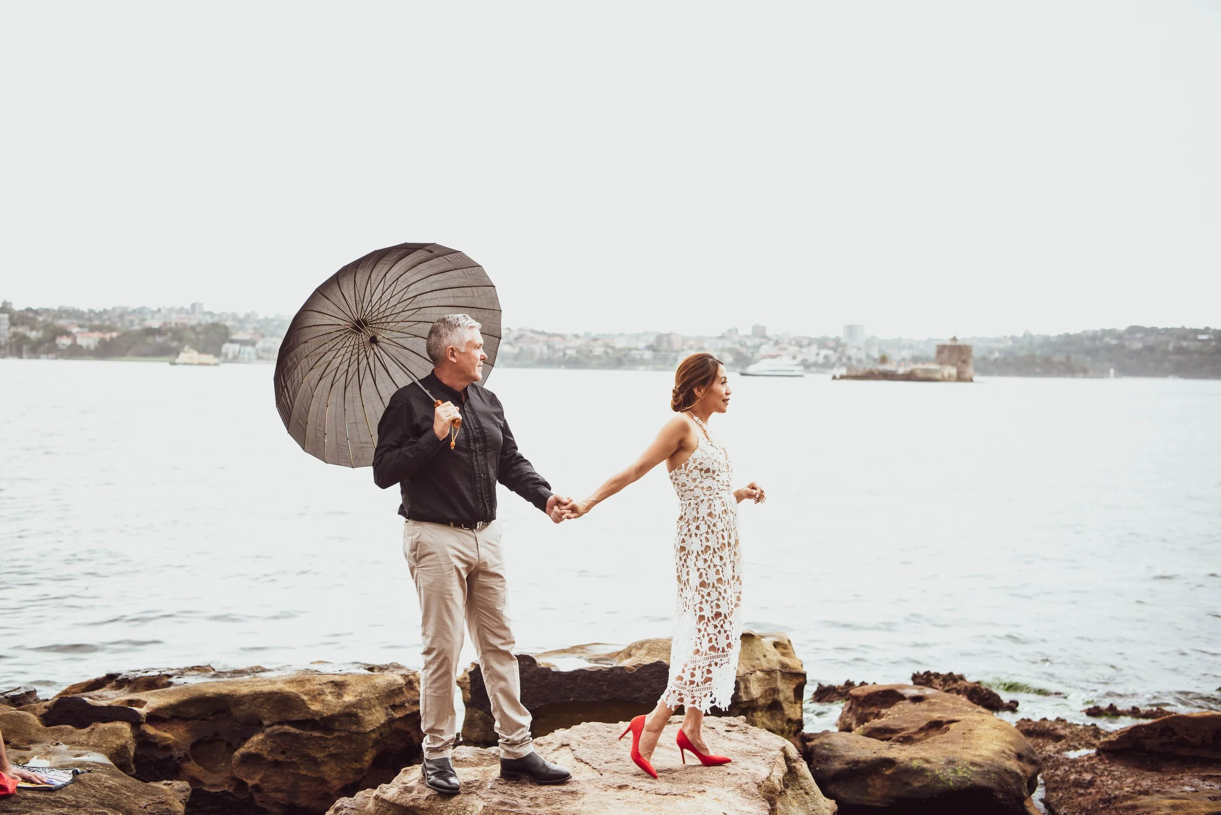 A couple stands on rocks by the water; the man holds an umbrella and is holding the woman's hand, who is wearing a white lace dress and red high heels, with an urban shoreline in the background.