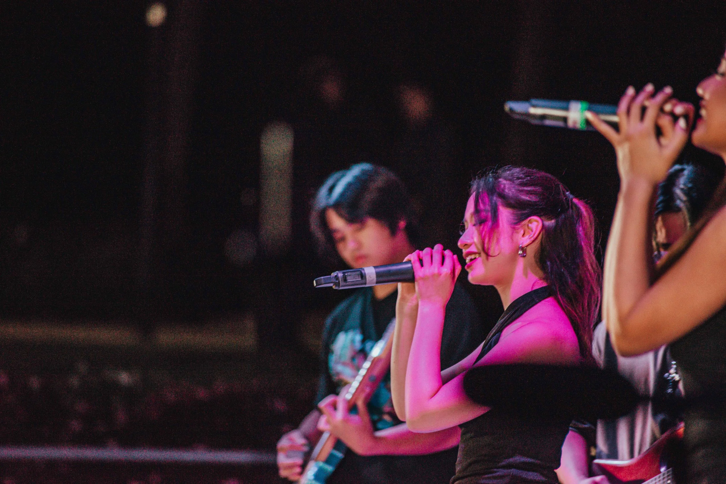A group of young women perform on stage, singing and playing guitars. The woman in the foreground holds a microphone and appears to be singing with her eyes closed. She has dark hair styled in a ponytail and is wearing a black top. The woman next to 