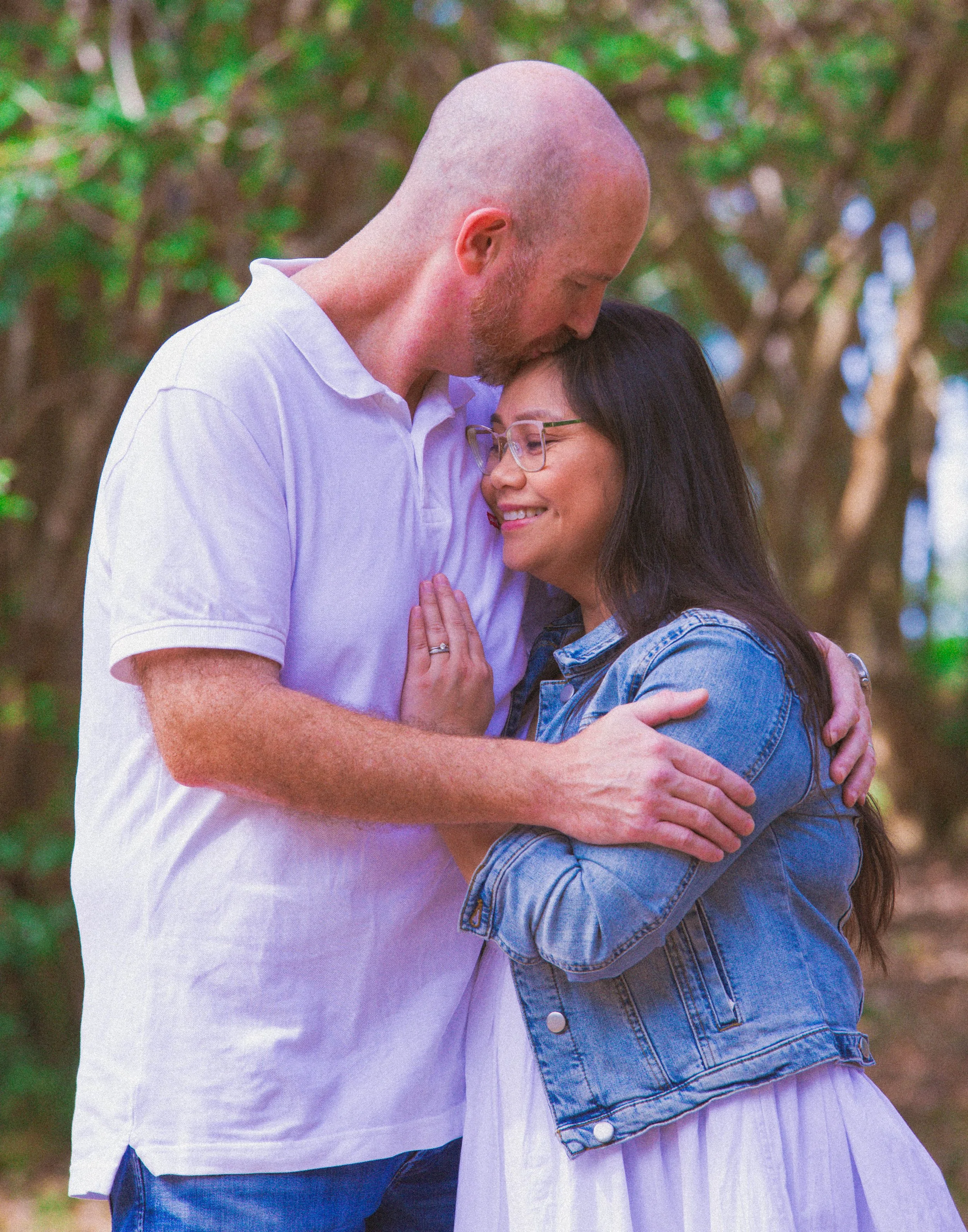 A man and a woman embrace outdoors, with the man gently resting his forehead on the woman's head, both smiling softly.
