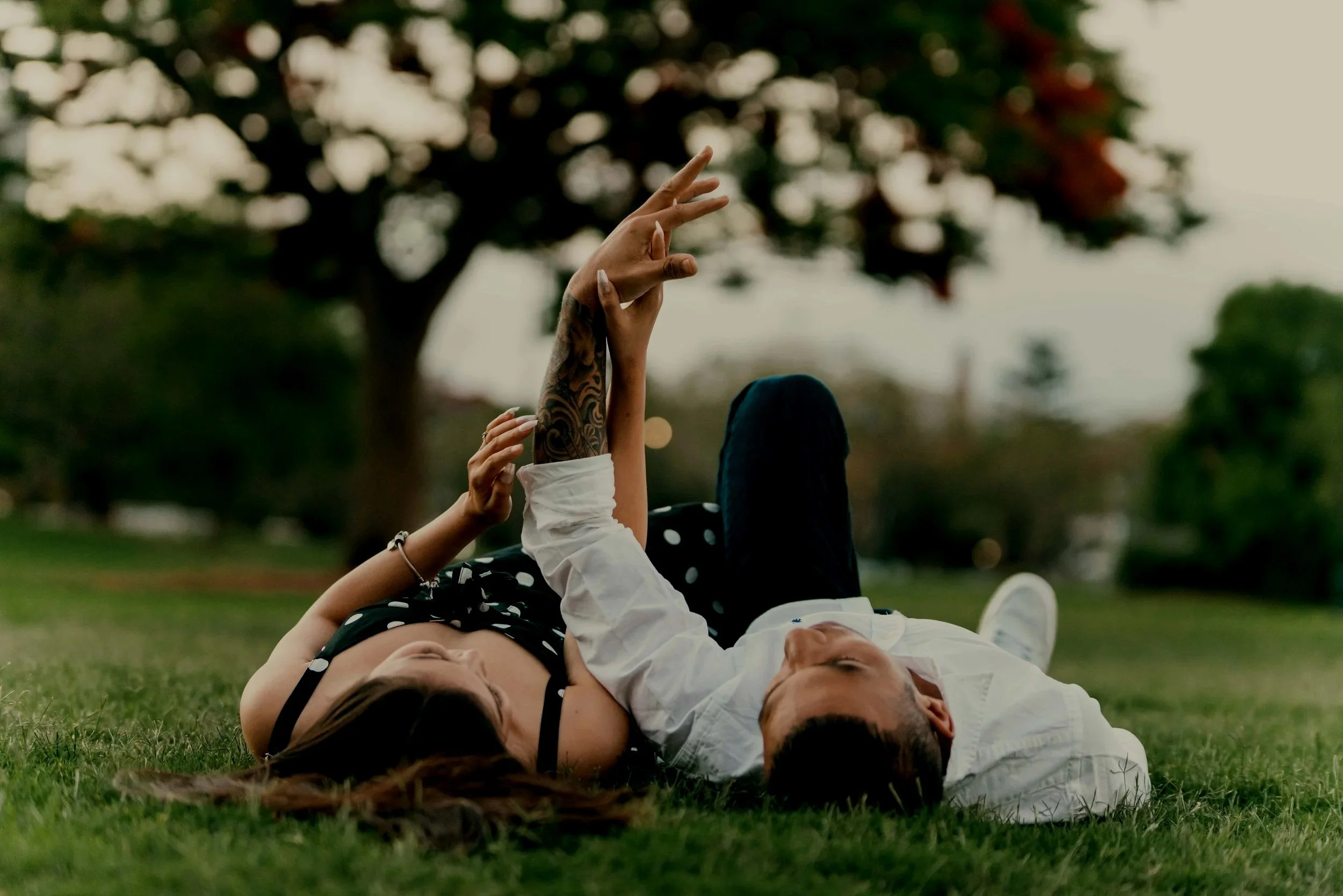A couple lying on the grass in a park, with the woman reaching up with her hand while the man looks at her, surrounded by trees and blurred background.