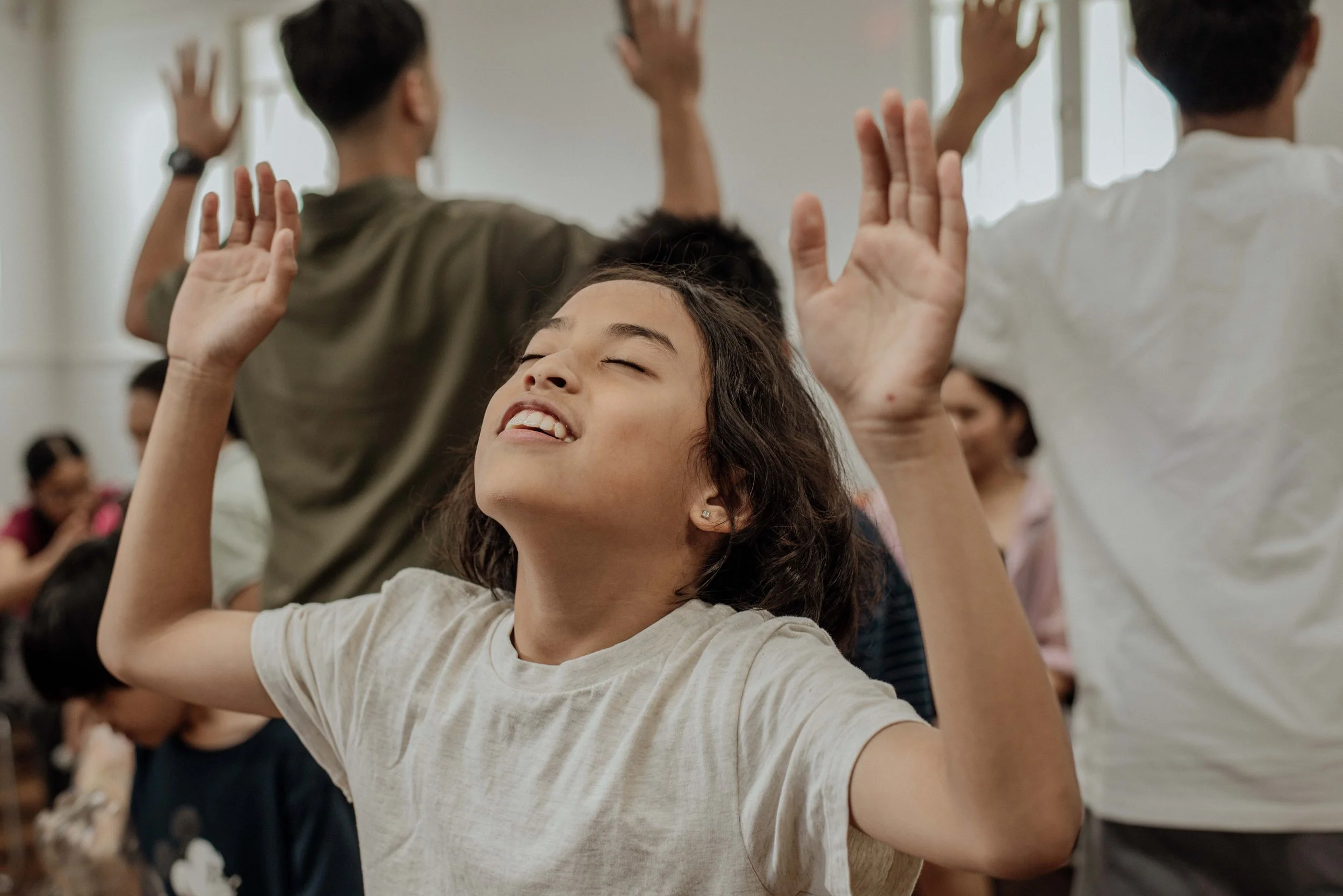 A girl with curly hair smiling and raising her hands during a group activity or dance in a classroom setting, with other children in the background.