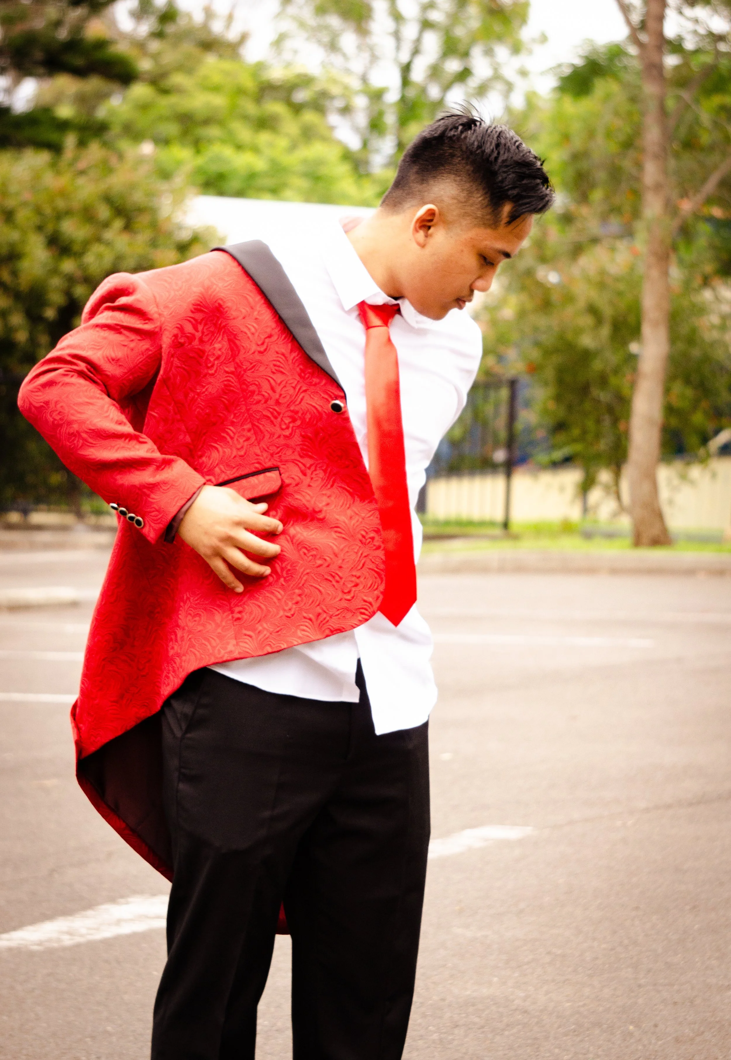Young man in formal attire, red blazer, white shirt, and red tie, standing in an outdoor parking lot looking down with hand on his stomach.