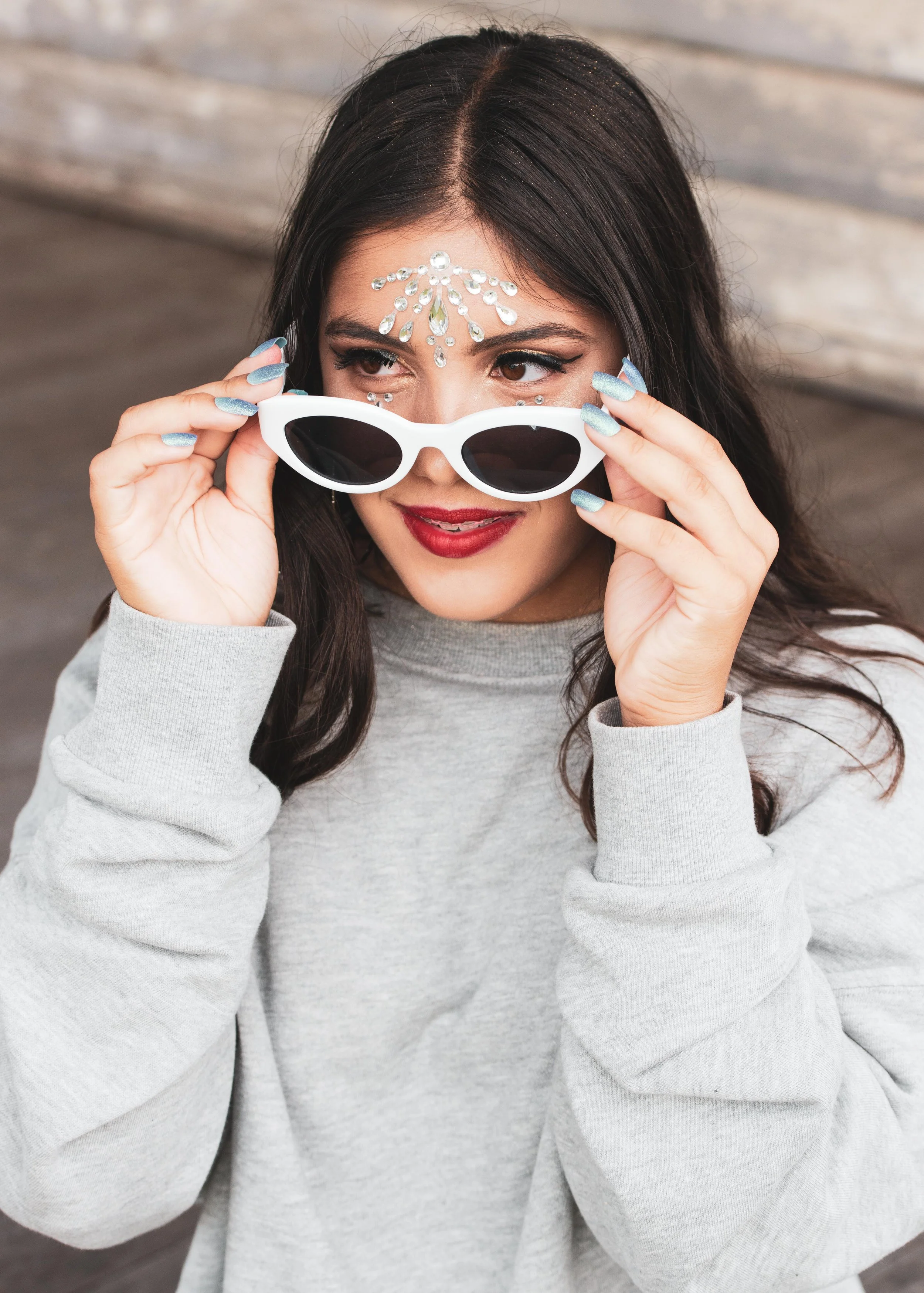 A woman with dark hair and red lipstick holding white sunglasses, wearing rhinestone face decorations, and a gray sweatshirt.