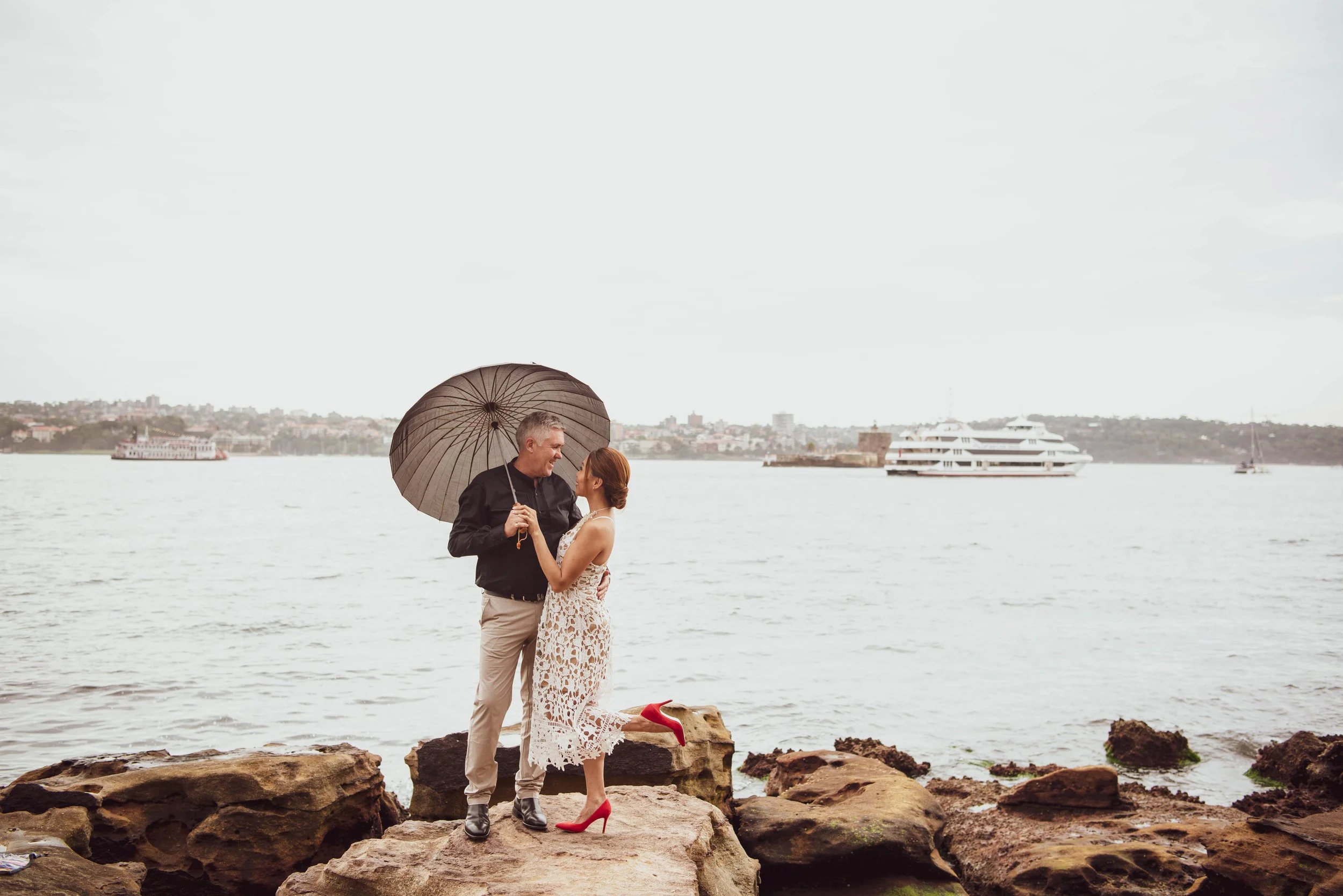A couple stands on rocks near water, sharing an umbrella. The woman wears a white lace dress with red high heels; the man wears a black jacket and beige pants. In the background, there is a large cruise ship and a cityscape across the water.