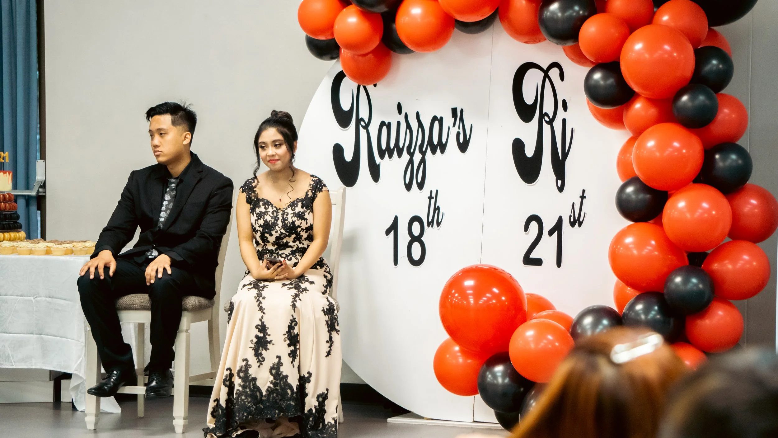 A man and a woman sitting on chairs at a birthday celebration, with black, orange, and red balloons and a sign reading 'Raizza's 18th 21st' in the background.
