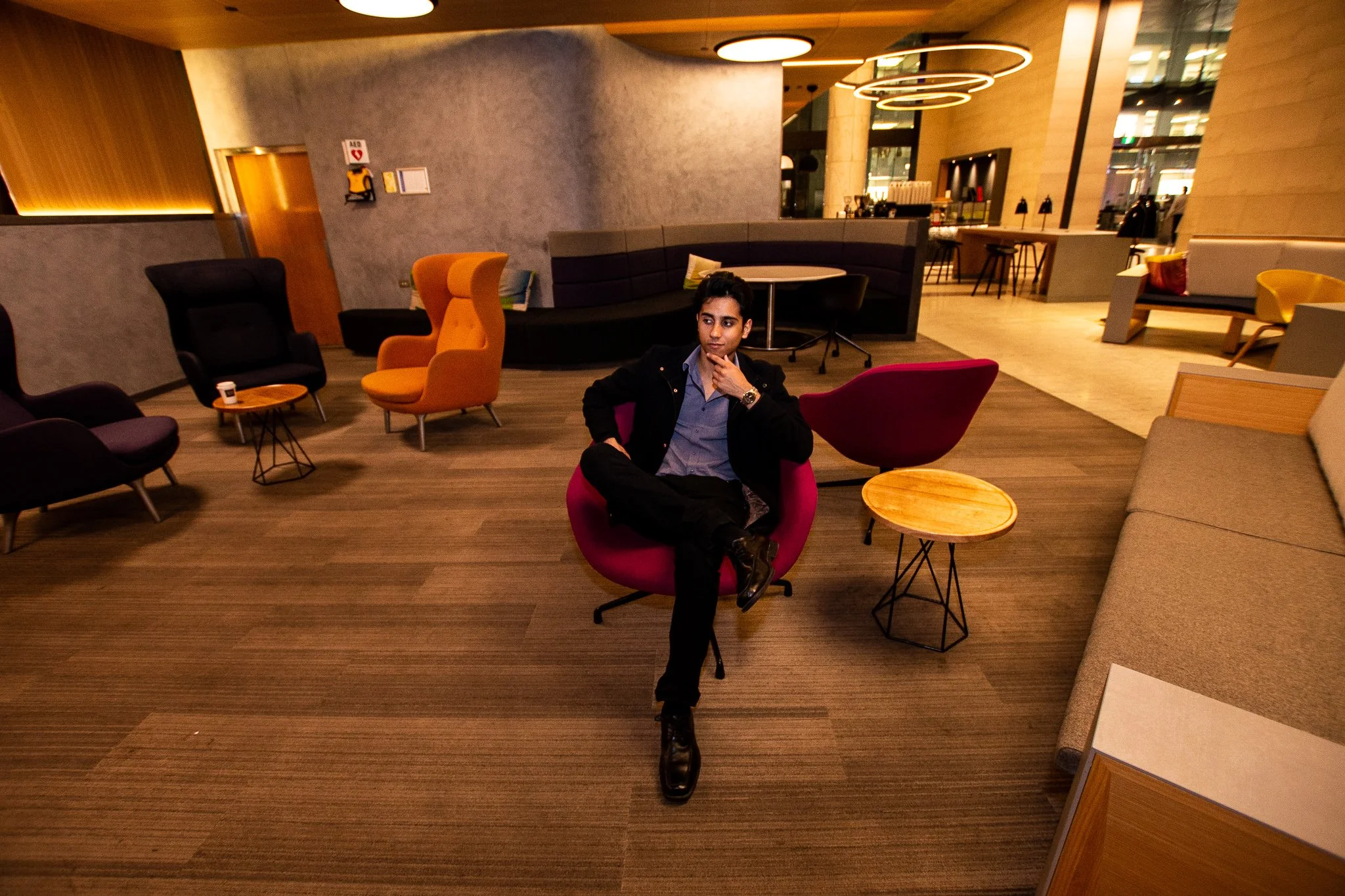 Young man sitting in a modern lounge area, resting with his legs crossed, surrounded by various contemporary chairs and tables, with a bar or café section visible in the background.