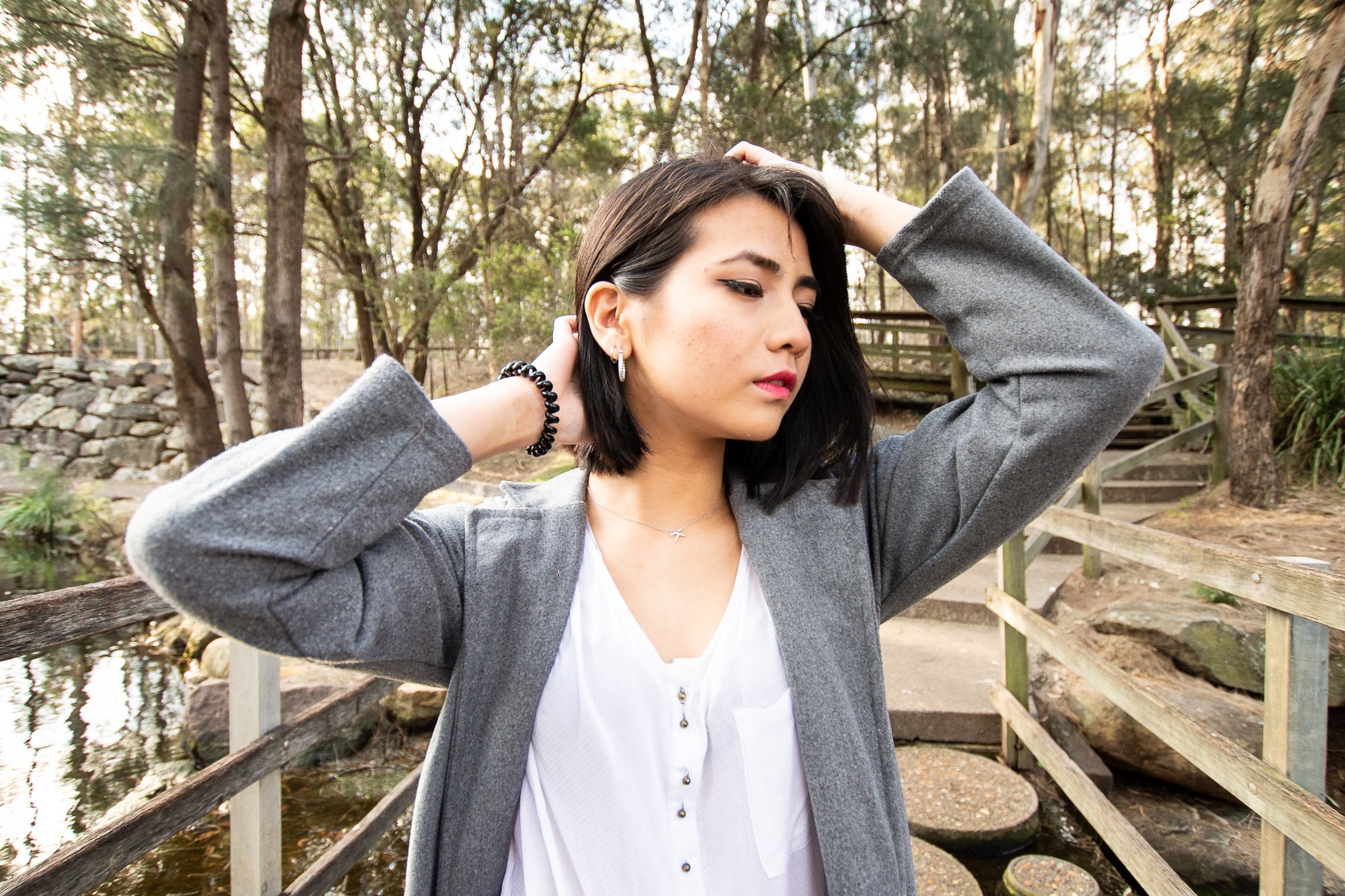 A young woman with dark hair, wearing a gray blazer and white top, fixing her hair in an outdoor park setting with trees, a pond, and wooden stairs.