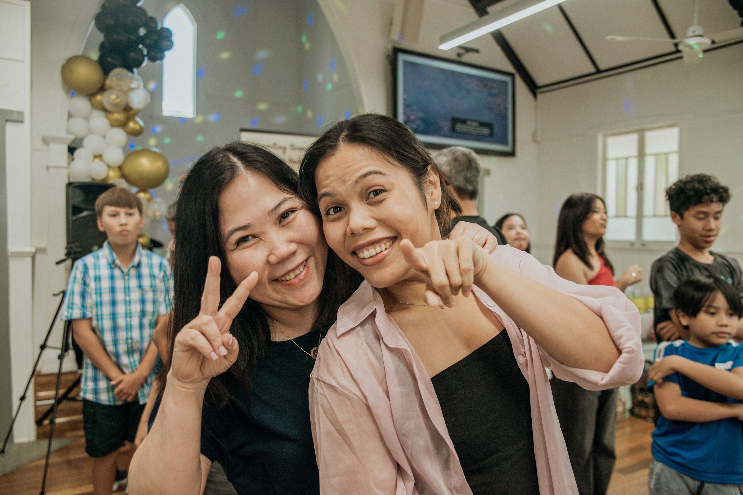 Two women smiling and making peace signs at a party with balloons and other guests in the background.