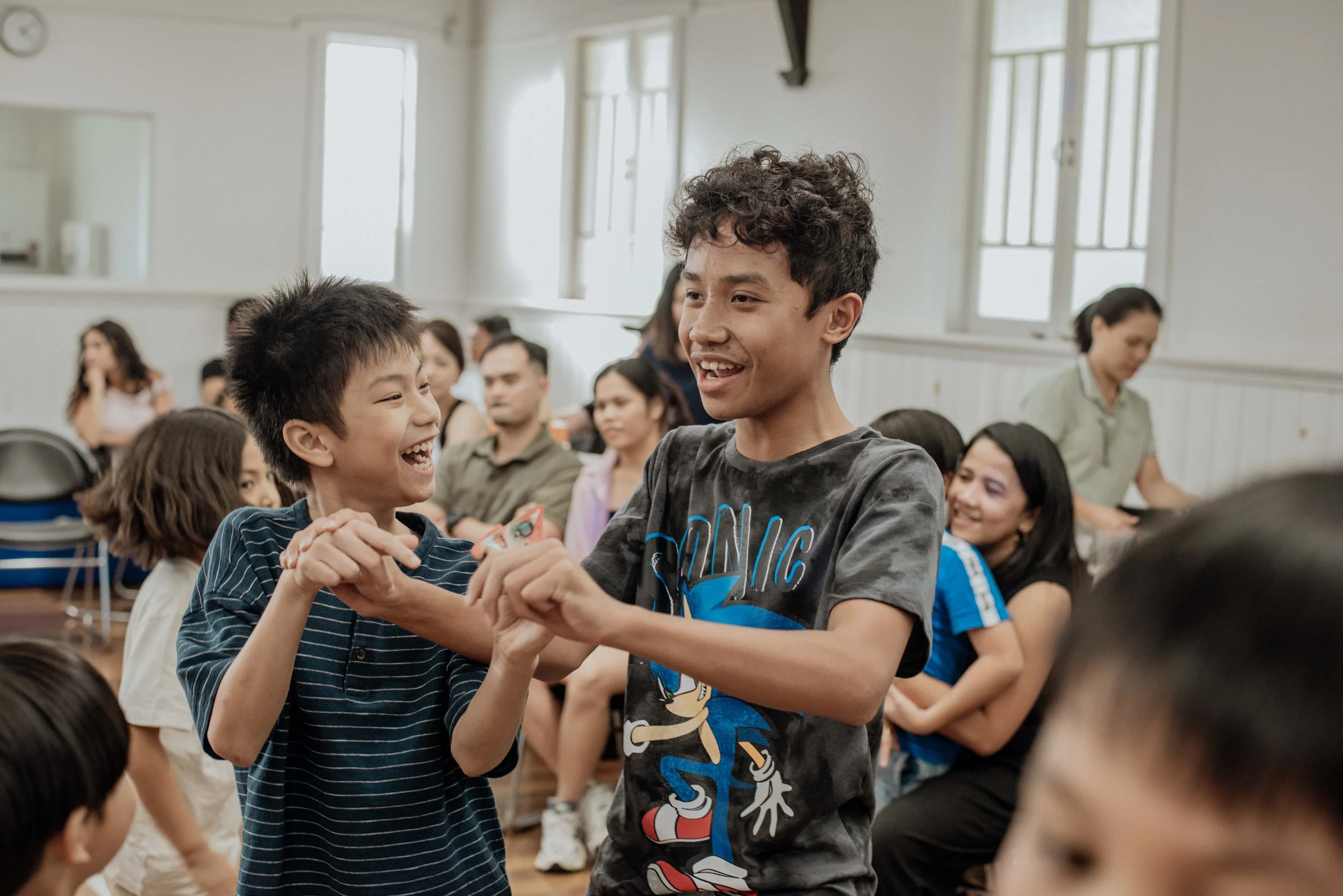 Two boys are happily dancing together in a room filled with children and adults, with a woman standing in the background.