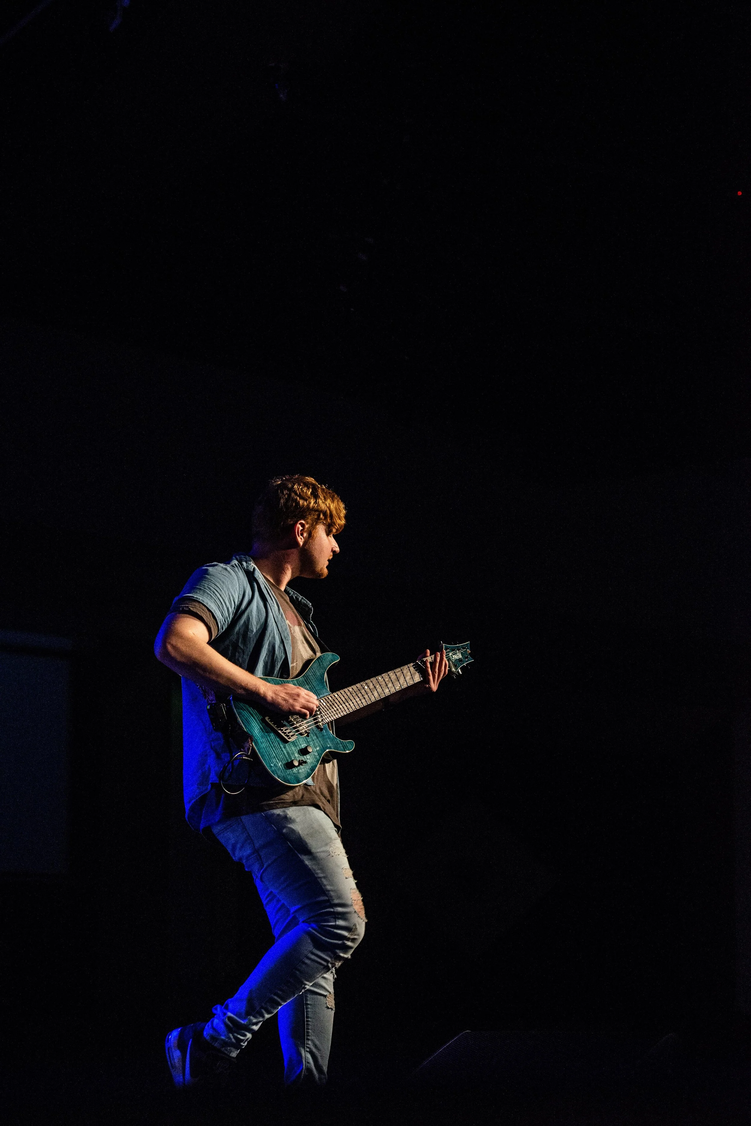 A young man playing an electric guitar on stage under a dark background, illuminated by stage lights.