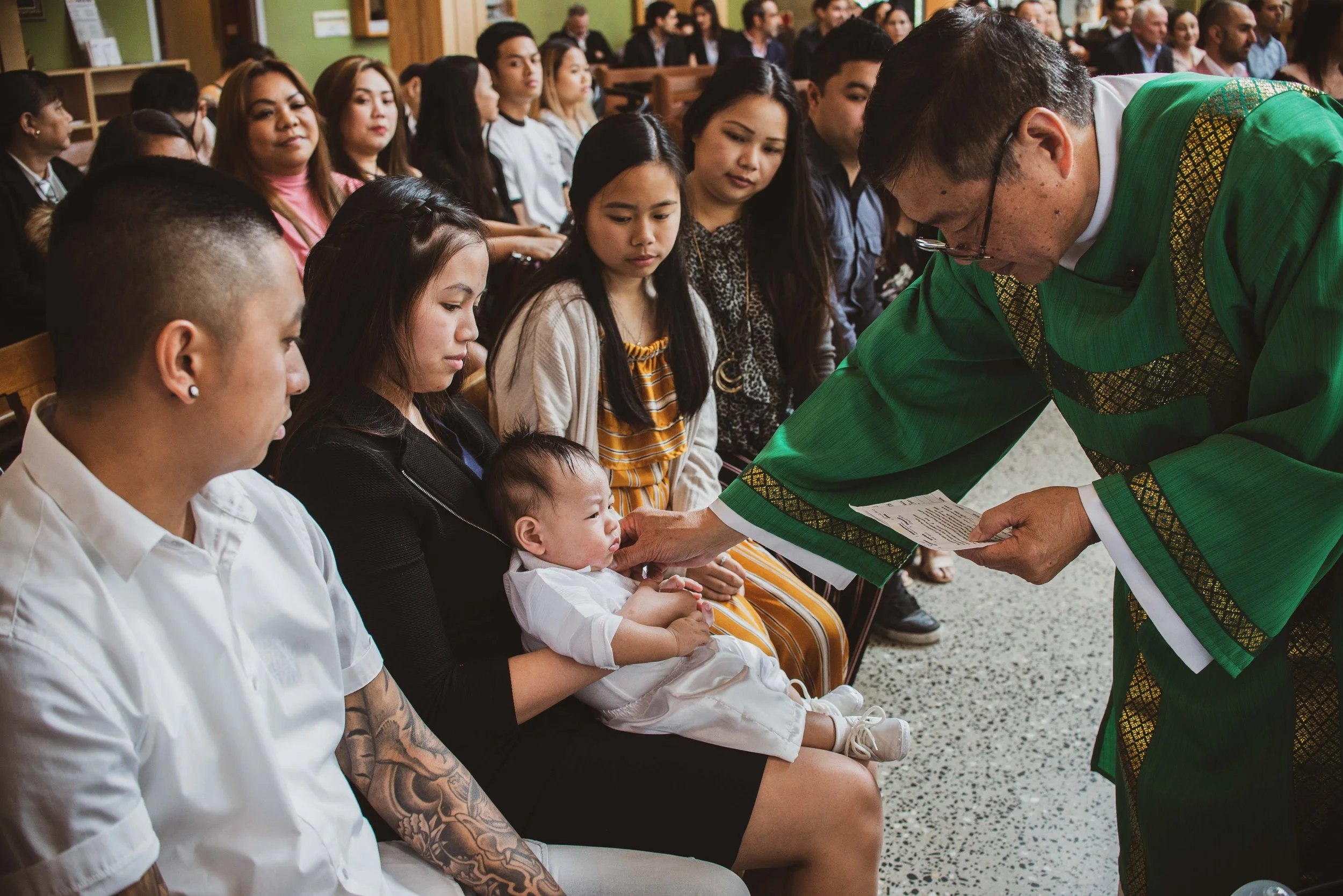 A religious ceremony, possibly a baptism or christening, taking place in a church. A priest in green robes is blessing a baby held by a woman. The woman is seated with other people, including a man with tattoos and a woman in a striped dress, surroun