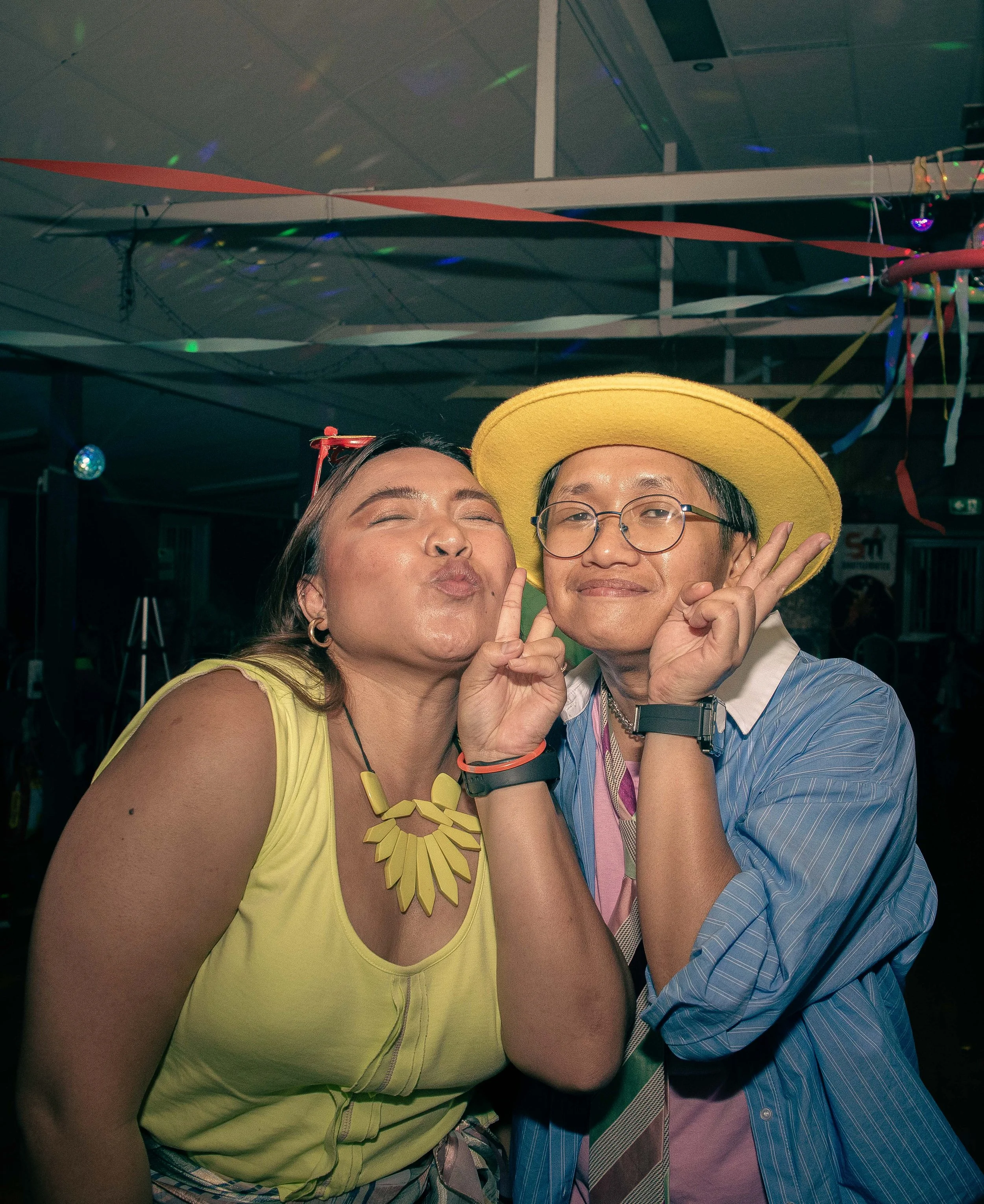 Two people dancing at a party, making peace signs and posing for the camera in a festive environment with colorful streamers and lights.