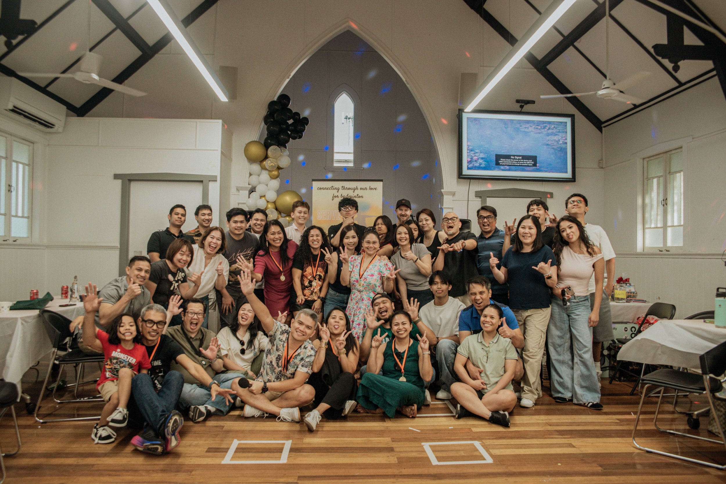 A large group of people smiling and posing together for a photo in a decorated indoor space, possibly a church or community hall, with balloons and a sign in the background that says 'connecting through our love for badminton'.