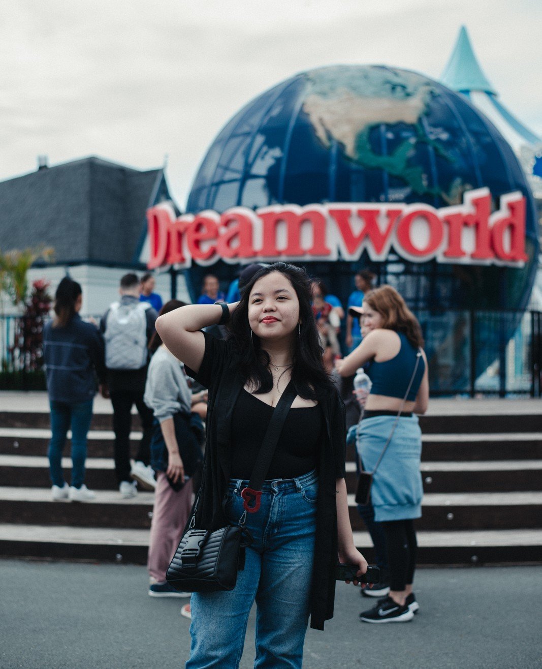 Young woman with black hair, wearing a black top and blue jeans, standing in front of the Dreamworld amusement park entrance with a large globe and the Dreamworld sign in the background.