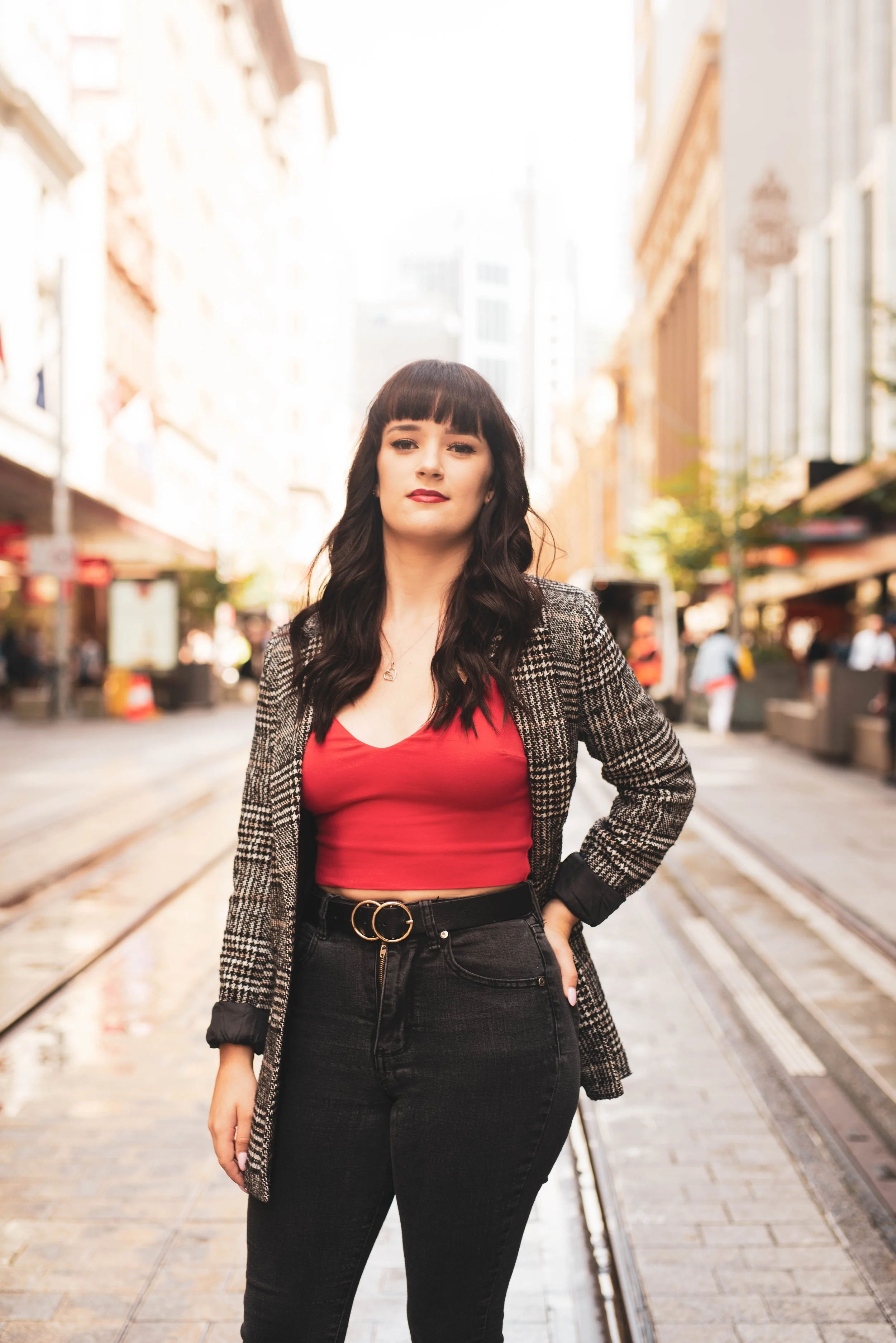 A woman with black hair and bangs standing on a city street with tram tracks, wearing a red top, black jeans with a belt, and a checkered blazer.