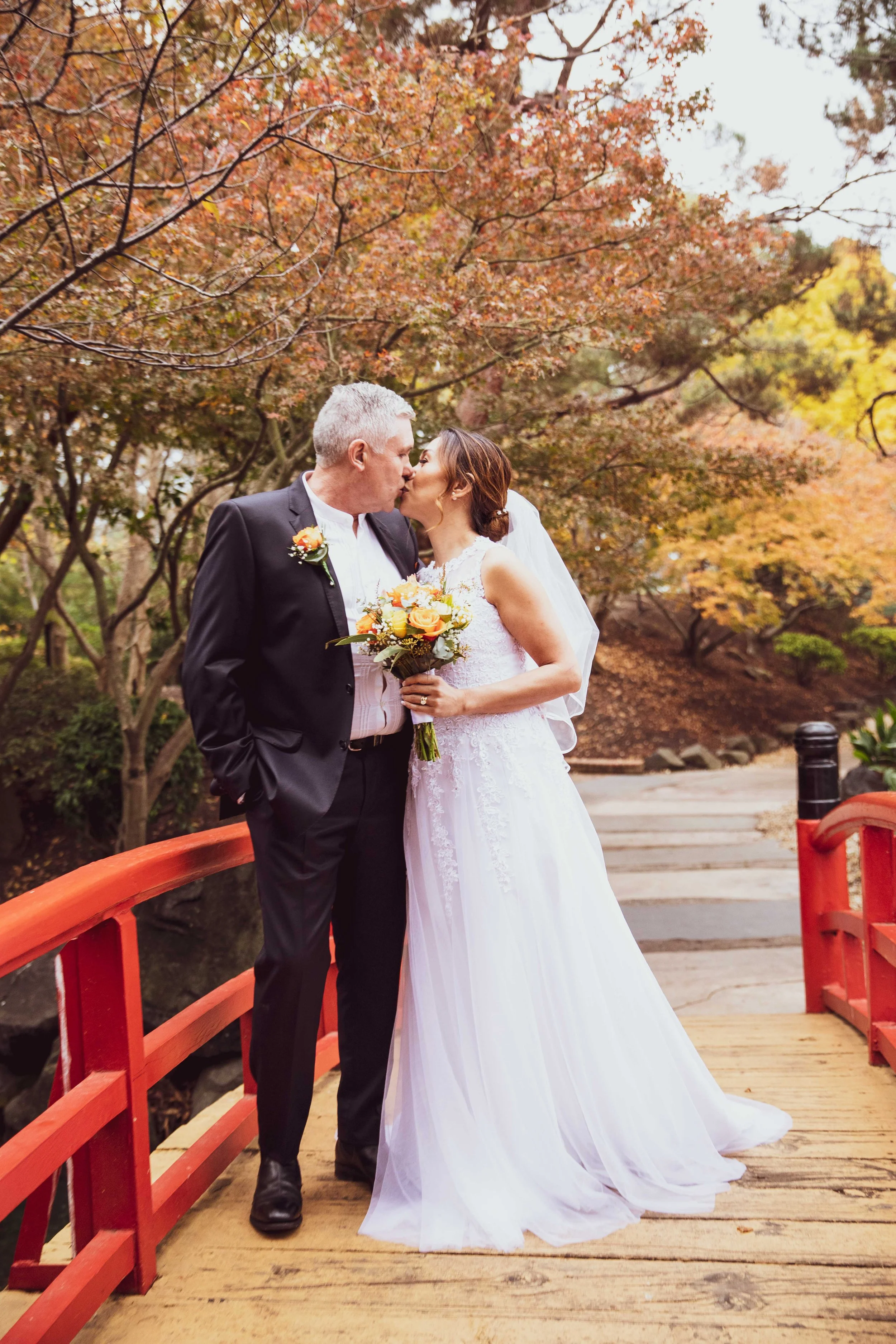 A bride and a groom share a kiss on a wooden bridge surrounded by autumn trees, with the bride holding a bouquet of orange, yellow, and white flowers.