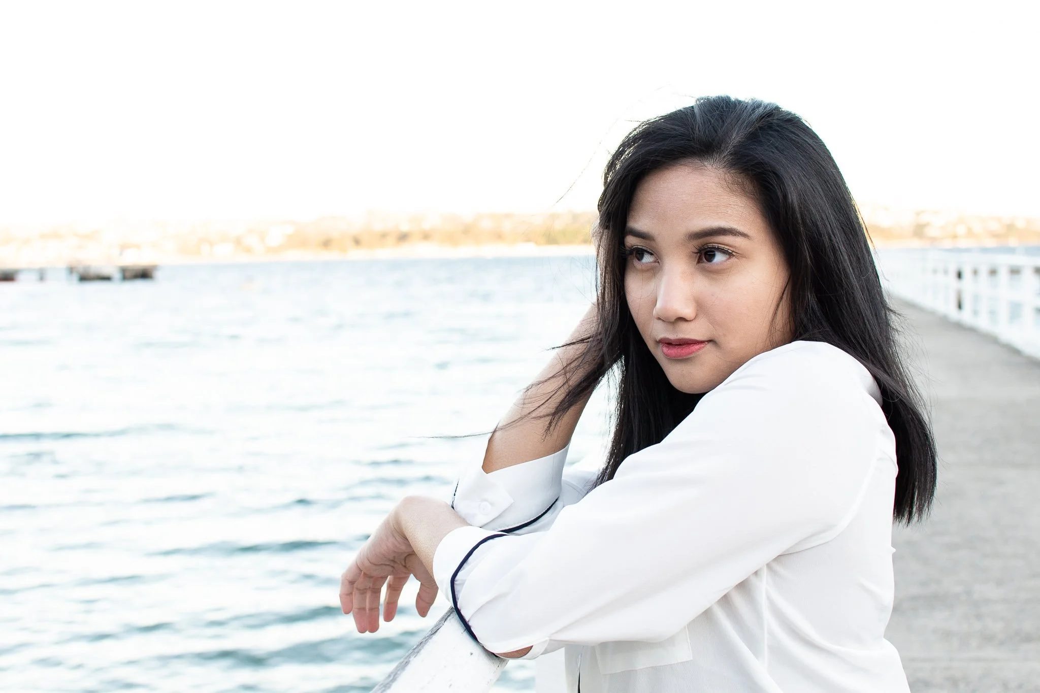 A young woman with black hair leaning on a railing by a body of water, looking to the side, during daytime.