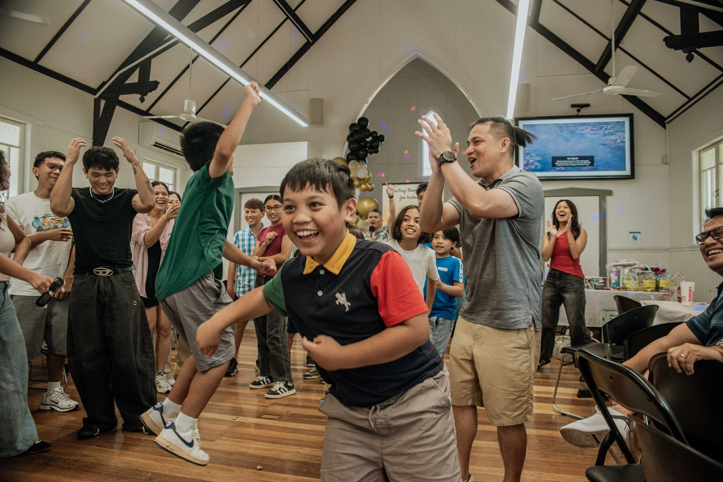 Children and adults dancing and celebrating in a decorated hall with balloons and a television screen.