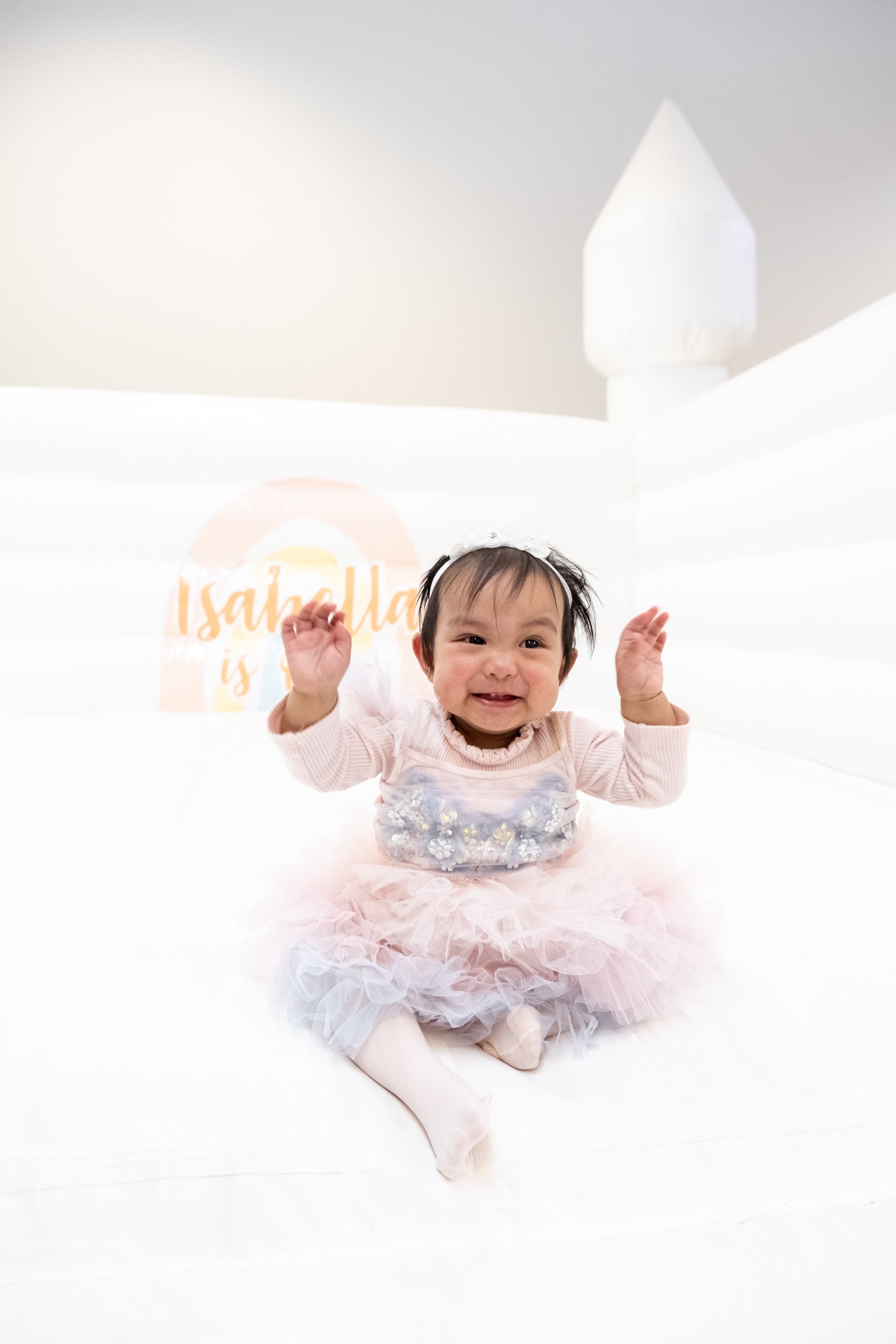 A smiling baby girl in a pink tutu dress sitting on a white surface, with a white background and a logo that reads "Isaiah is" in the distance.