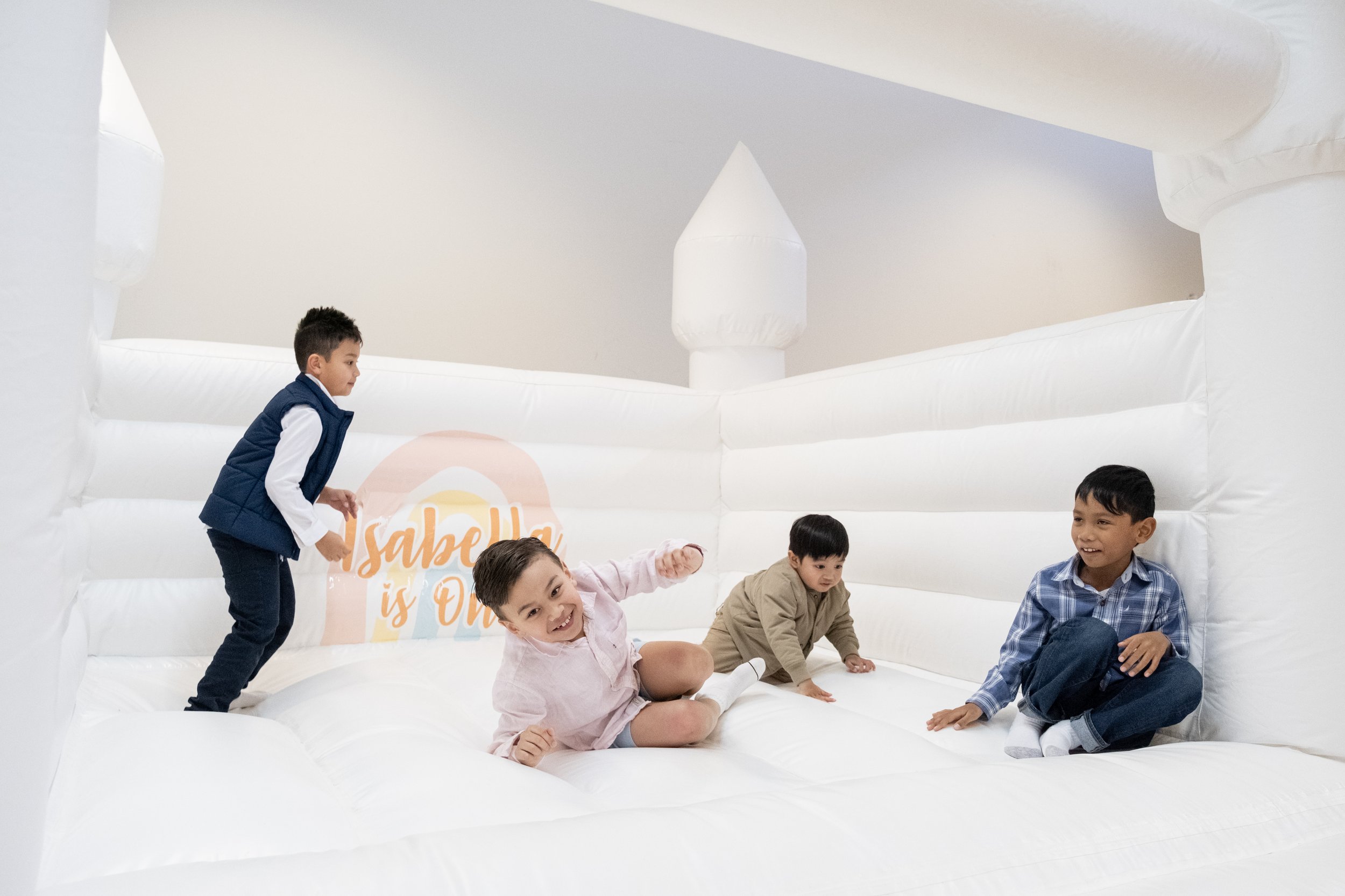 Four children playing inside a white inflatable bounce house with a sign that says "Isabelle is One".
