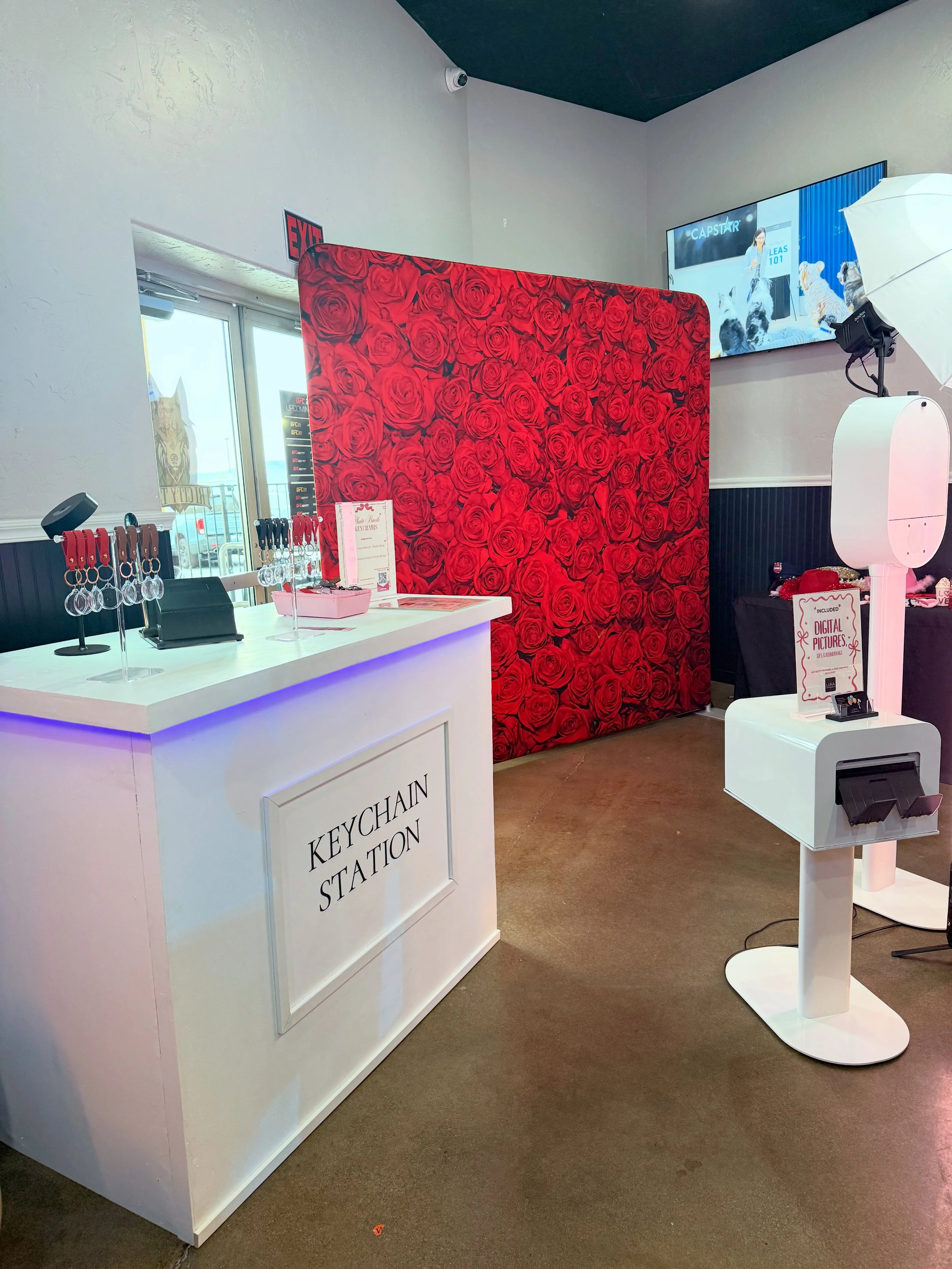 Elegant white keychain station setup with acrylic photo keychains on display rack next to open air photo booth with red rose backdrop by Luna Event Rentals in Tri-Cities WA