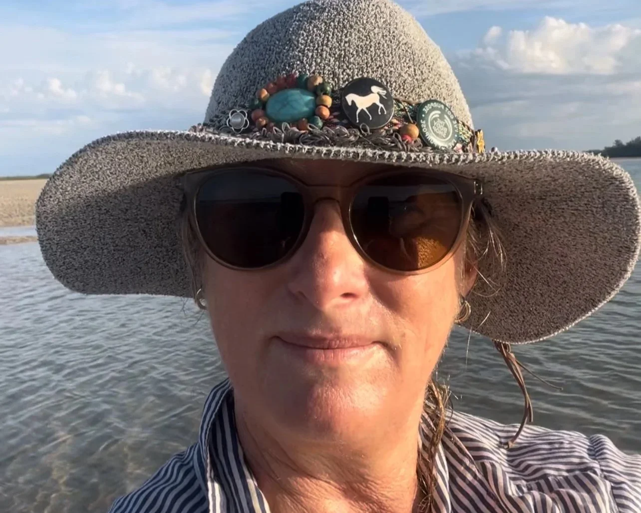 A face of a woman over 50 with a hat on and sunglasses and ocean in the background