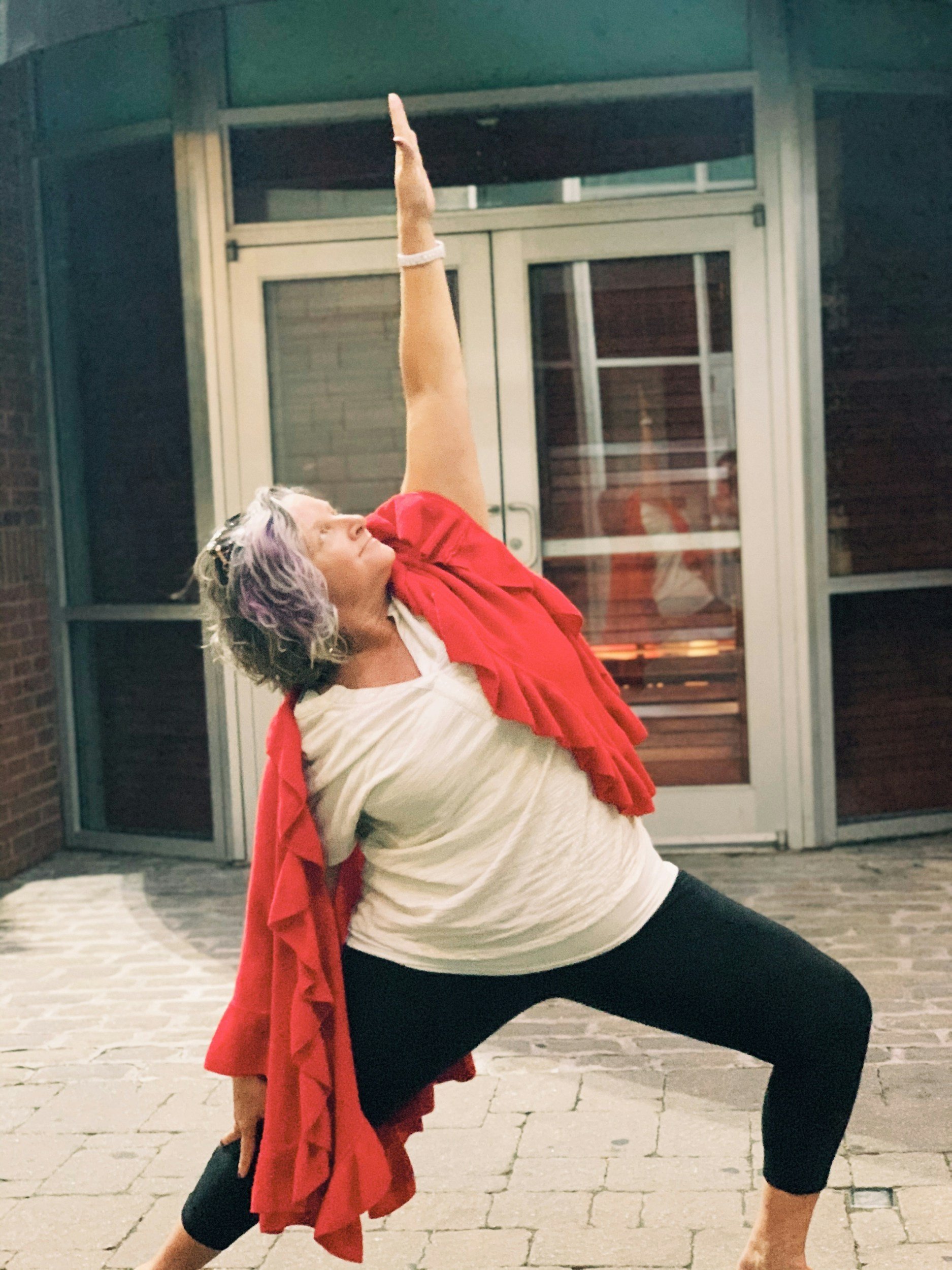 A woman with curly, short, gray hair practicing yoga outdoors on a paved area, striking a Warrior pose with one arm reaching upwards and the other arm stretched back, wearing a white t-shirt, black leggings, and a red jacket draped over her shoulders.