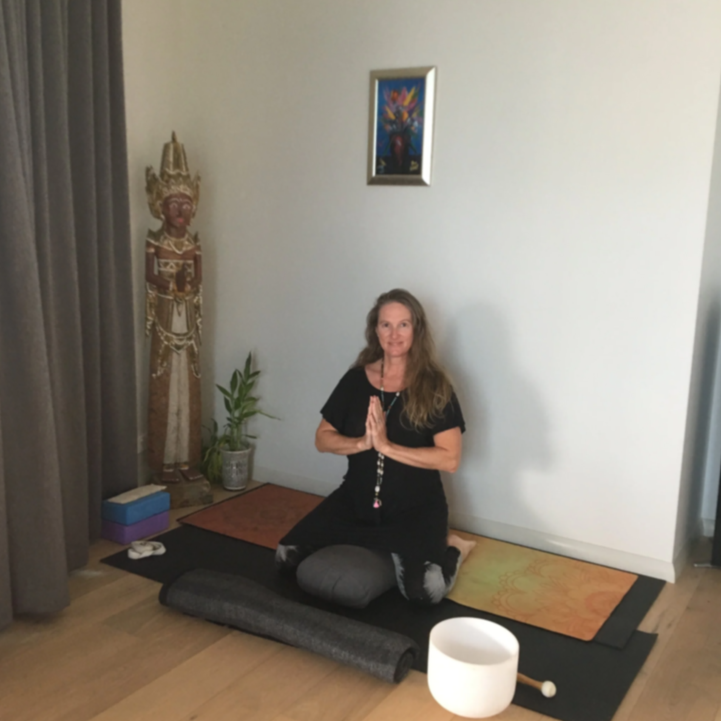 Woman practicing yoga in a meditation pose on a yoga mat in a room with spiritual decorations, including a statue and framed artwork.