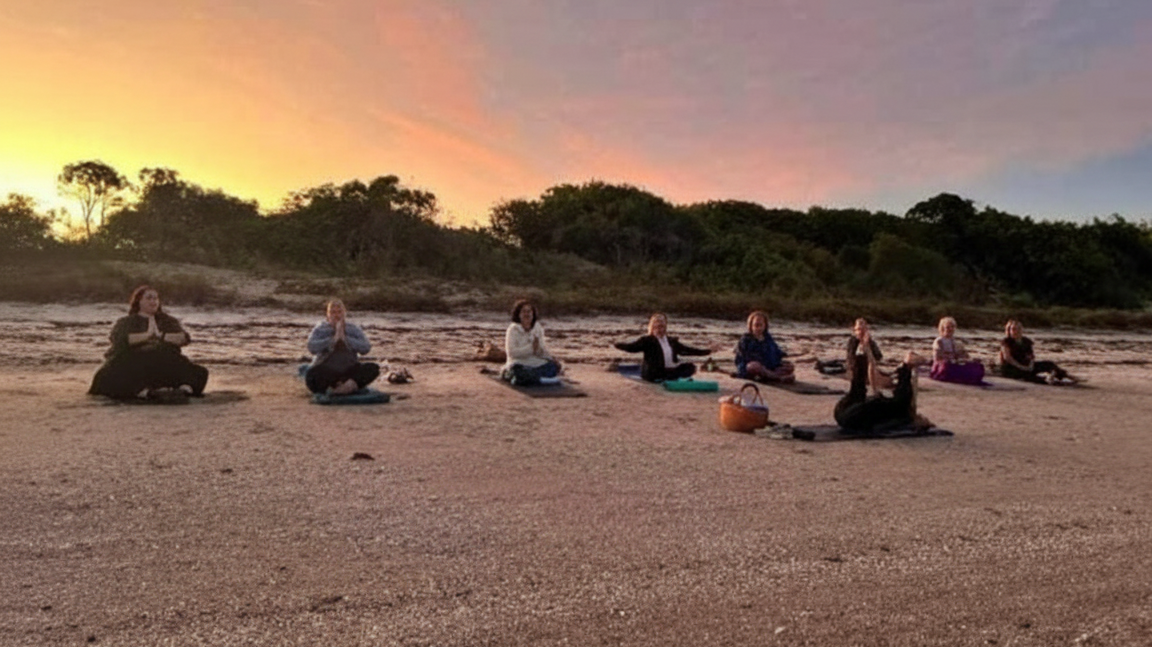 womens group practicing yoga on a beach during sunset, sitting on yoga mats in a row facing the ocean, with trees and colorful sunset in the background.