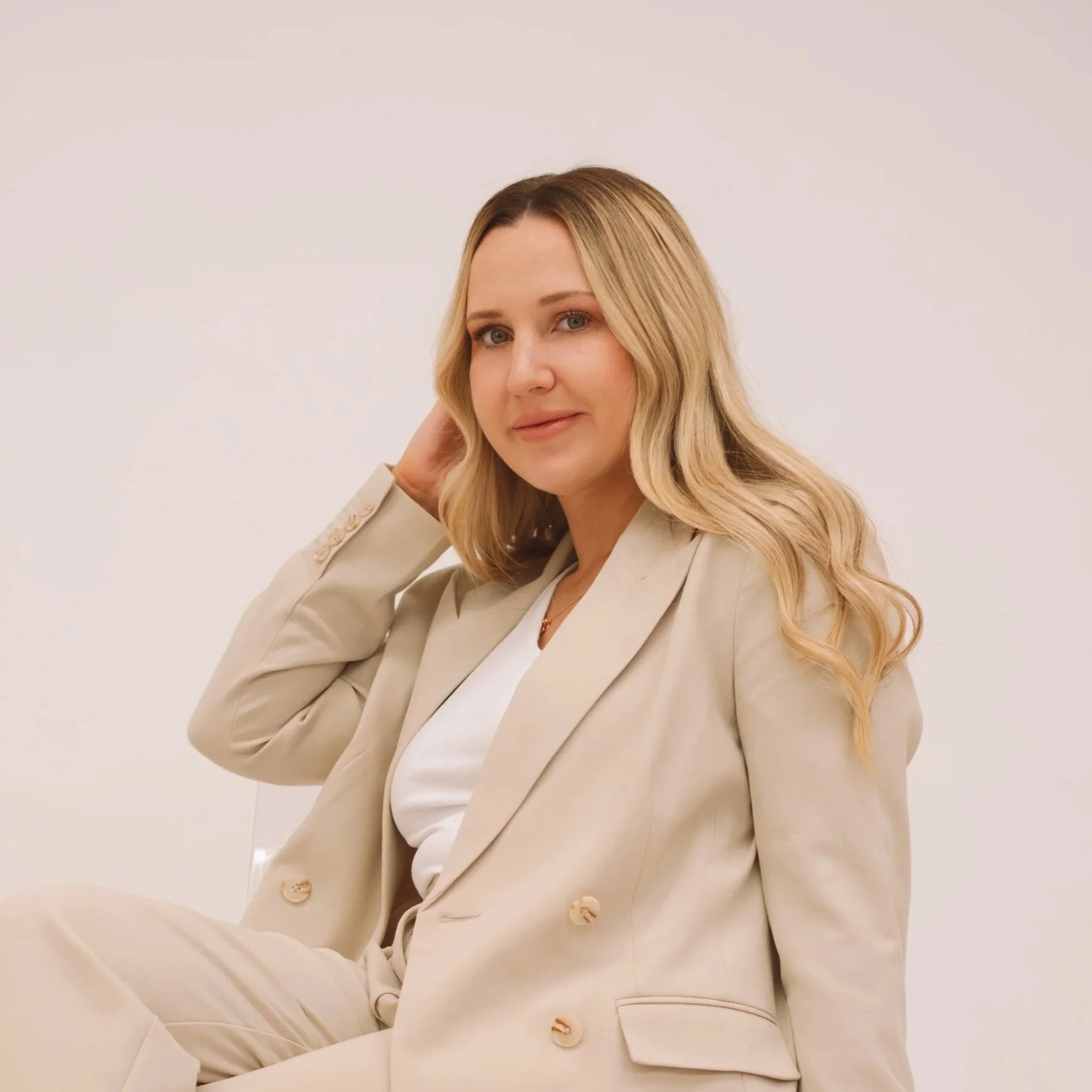 A woman with blonde wavy hair wearing a beige suit sitting against a plain white background.