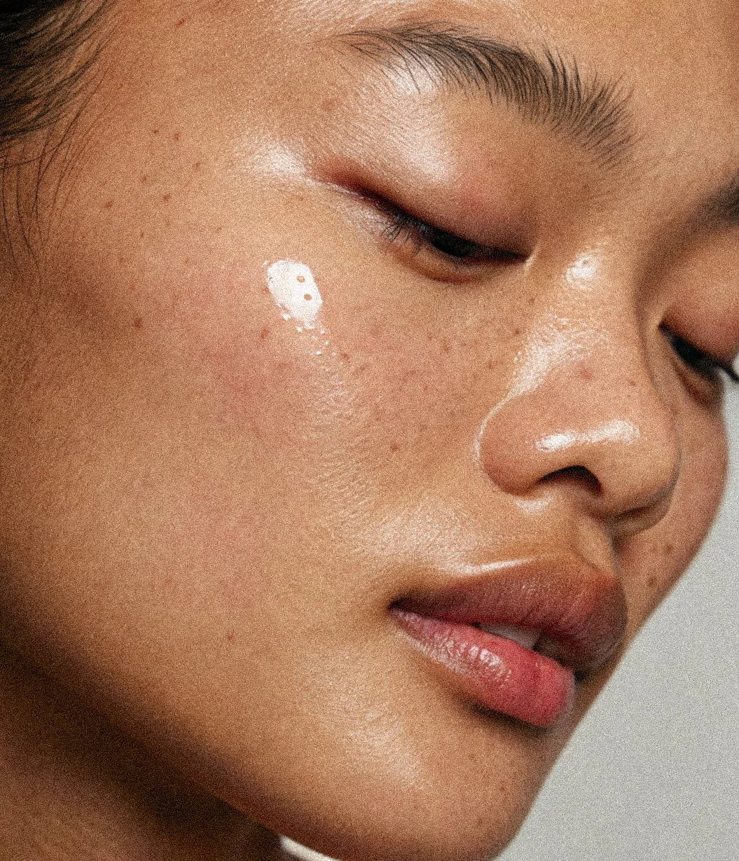 Close-up of a woman's face with smooth skin, showing her closed eye, nose, and lips, with a small amount of white cream on her cheek.