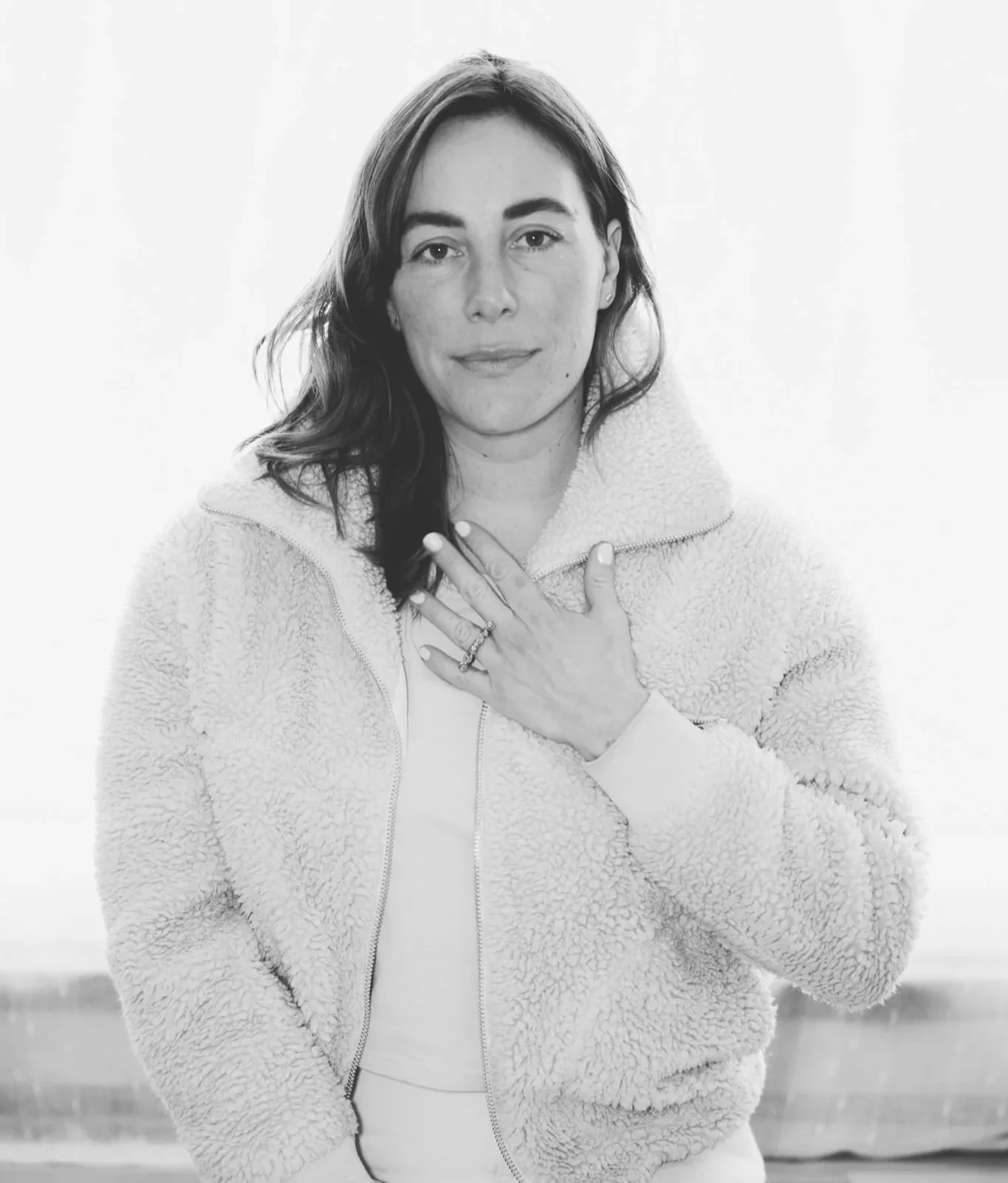 A black and white portrait of a woman with shoulder-length hair wearing a fluffy zip-up jacket, her left hand resting on her chest with visible rings.