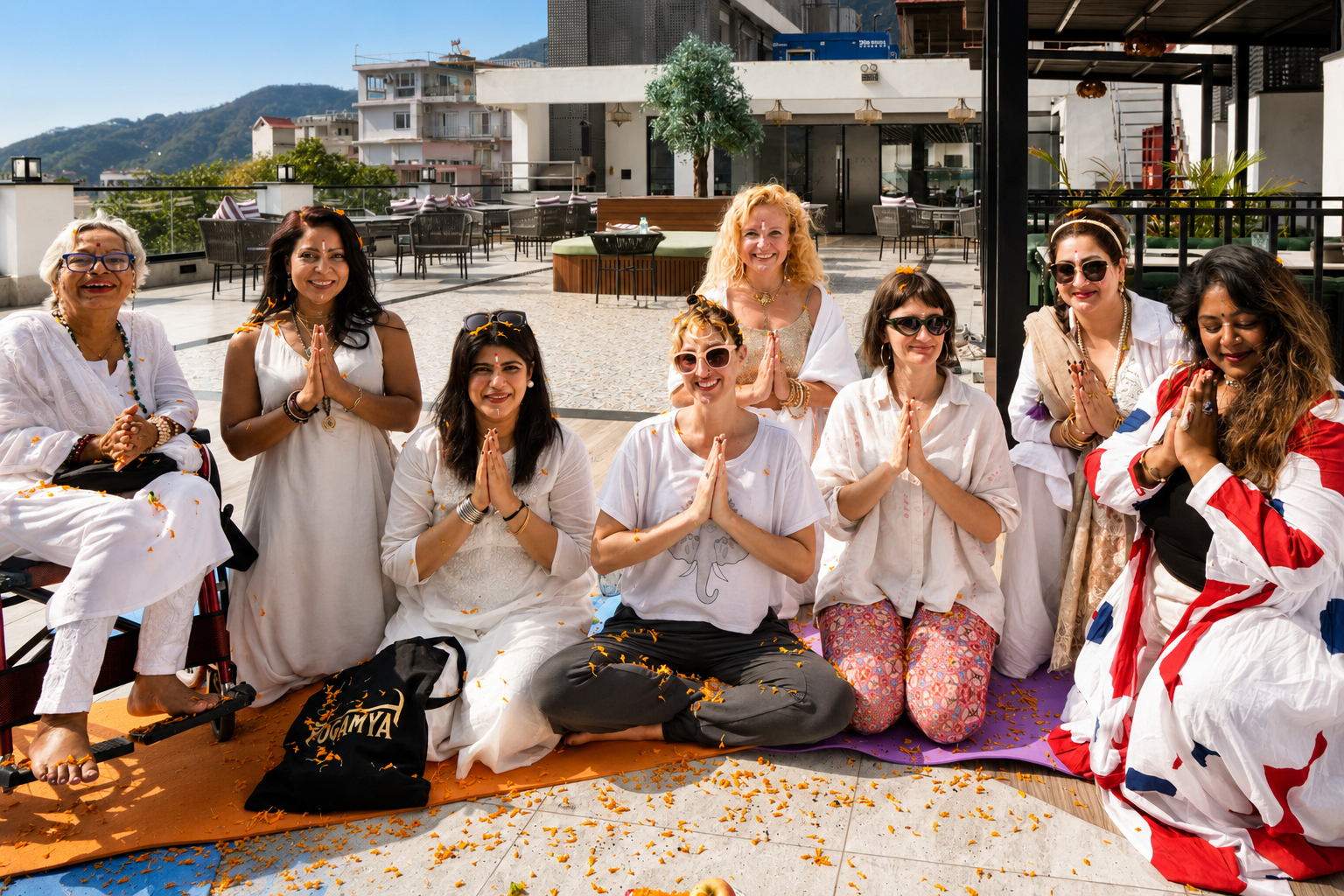 Group of nine women participating in a prayer or meditation session outdoors, sitting or kneeling on mats, with orange flower petals scattered on the ground, on a rooftop terrace with a cityscape and mountains in the background.
