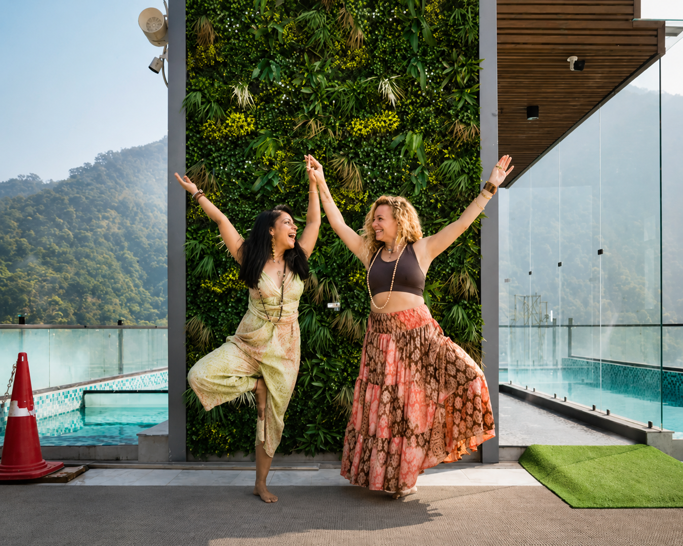Two women happily dancing and giving high fives in front of a green wall at a rooftop pool area, with mountains in the background.