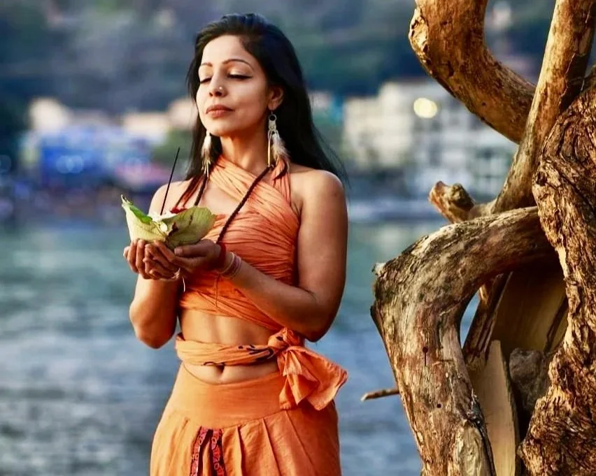 A woman in an orange wrap-around dress holds a green chameleon near a tree by a body of water, with a blurred cityscape in the background.