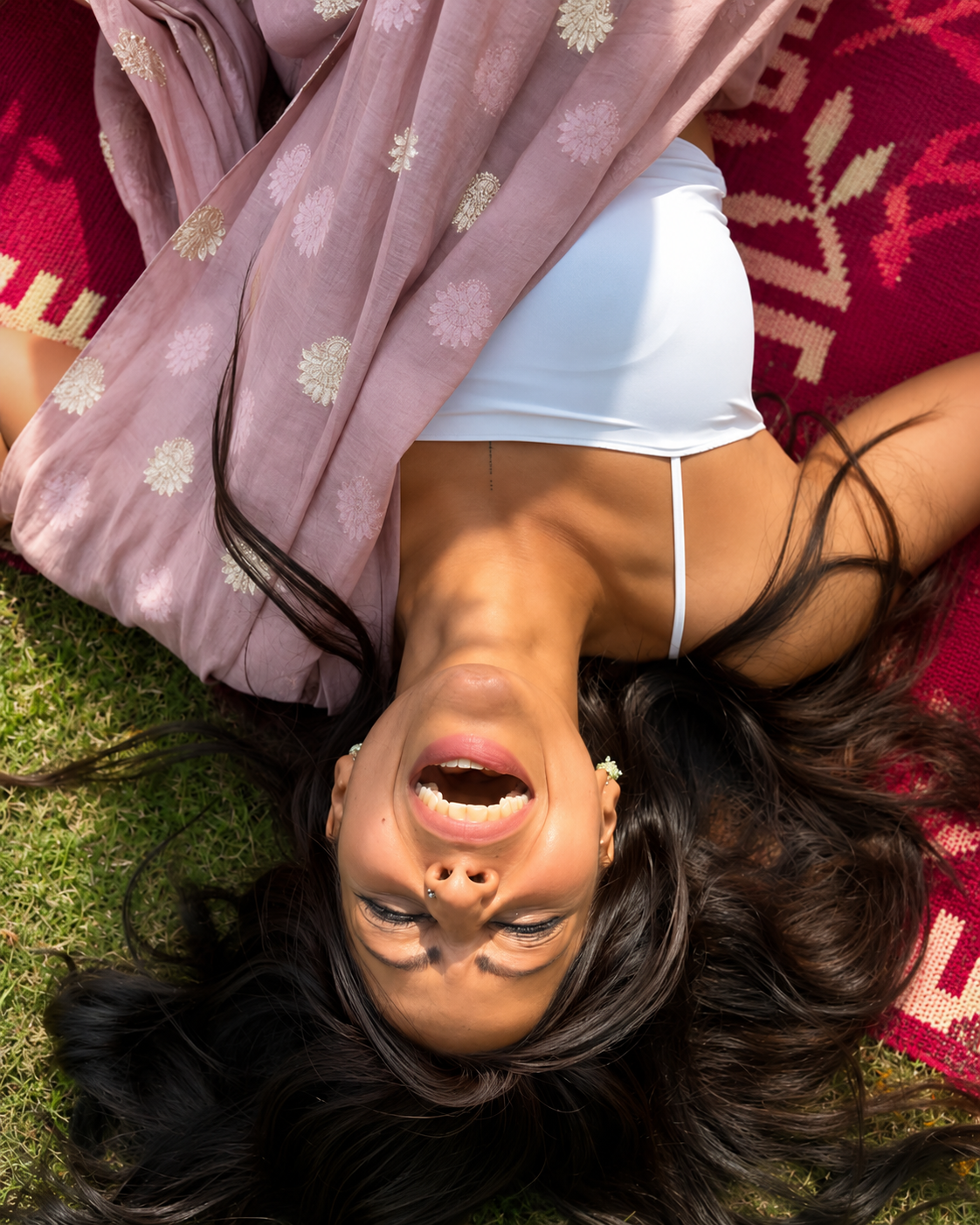 A woman lying on her back on grass with long dark hair spread around her, smiling and laughing, wearing a white top and pink scarf with floral patterns.