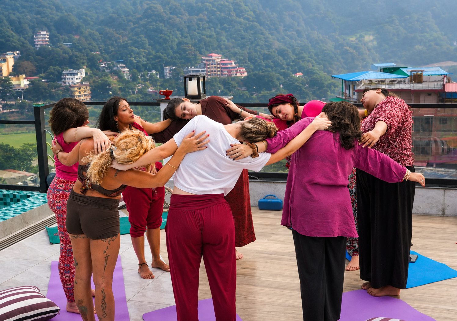 Group of women practicing yoga or meditation on a rooftop deck with a mountain view, some with eyes closed and others touching each other's shoulders or arms.