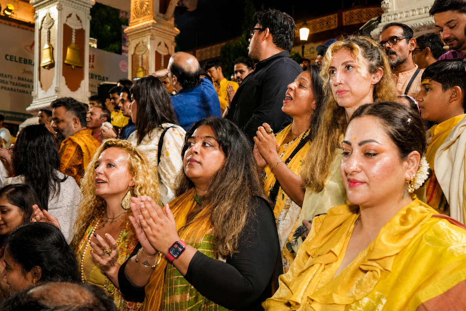 A group of people, including women and men, gathered at a religious or cultural event, many wearing traditional yellow and white garments, some with flowers and jewelry, showing expressions of devotion and reverence.