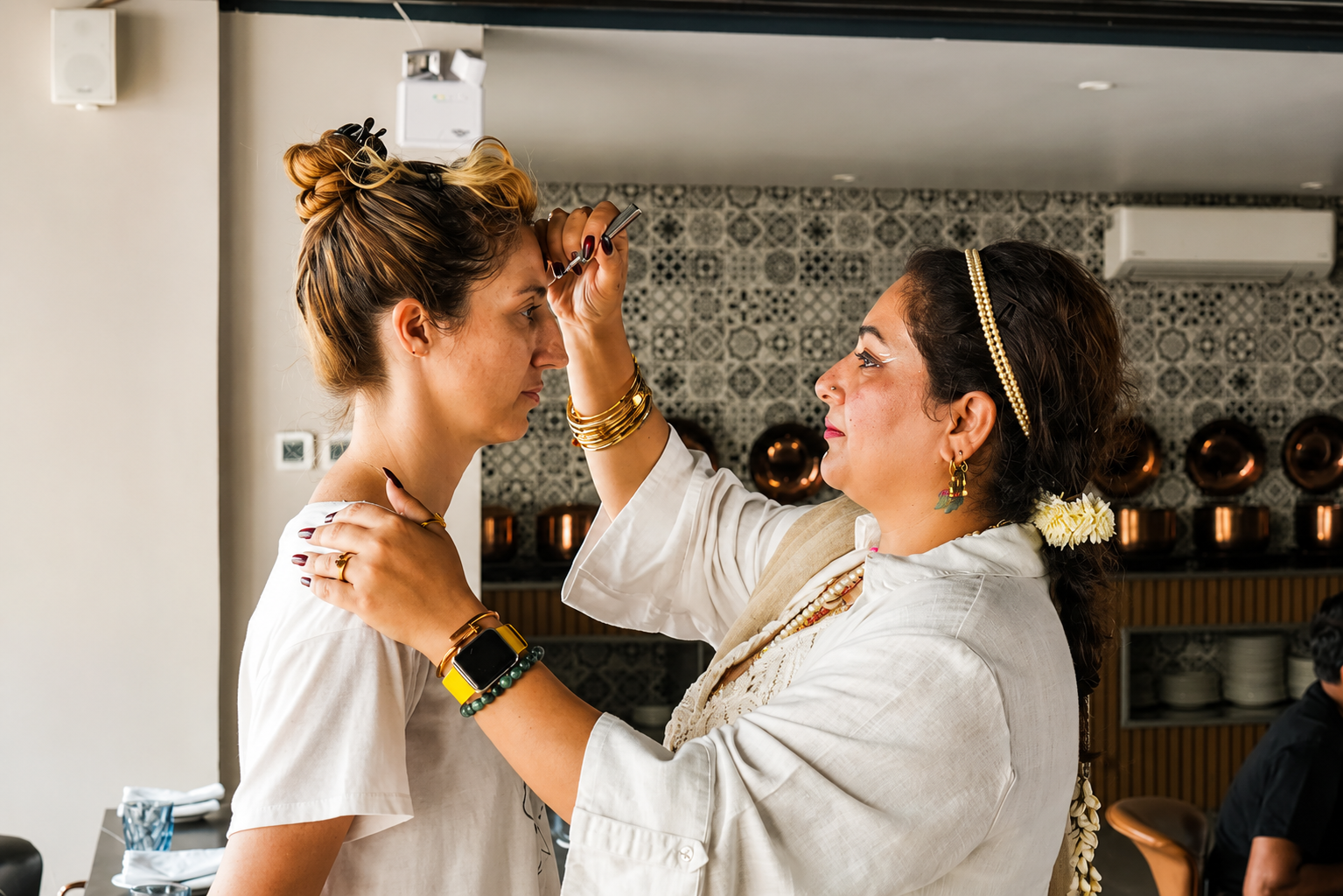 A woman with dark hair and colorful earrings is applying makeup to a younger woman, both dressed in white. The setting appears to be indoors with a patterned black-and-white wall and copper pots in the background.