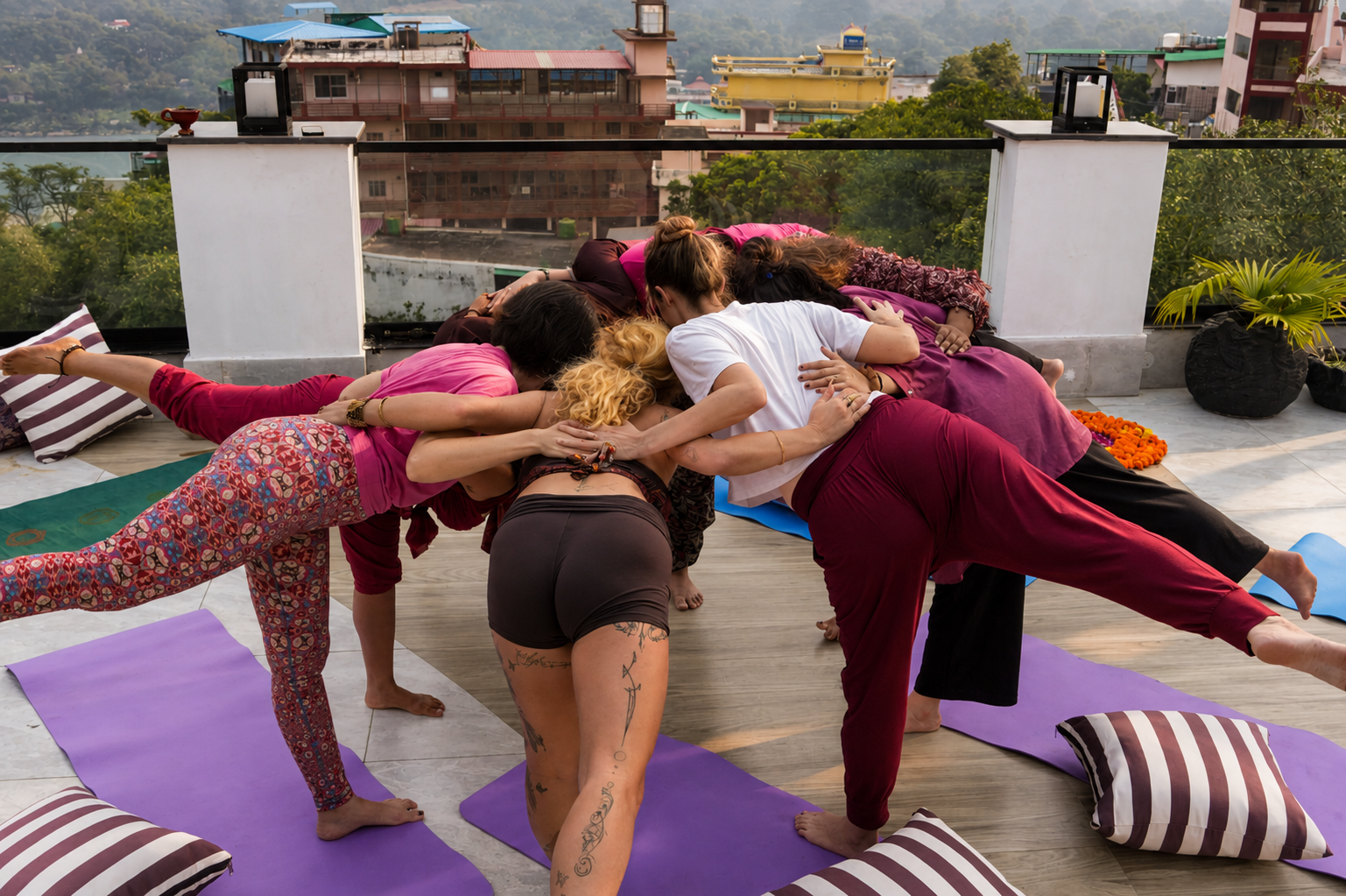 Group of people practicing yoga outdoors on a rooftop terrace, performing a partner pose, with colorful mats and pillows.