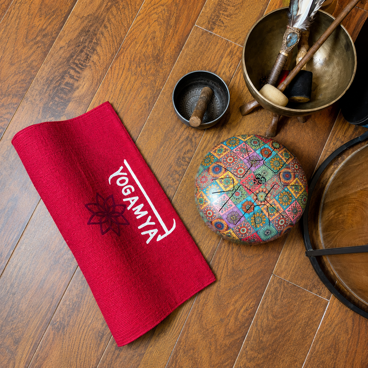 A red yoga mat, a colorful mandala-patterned meditation cushion, a singing bowl with mallets, a small black bowl, and wooden blocks are arranged on a wooden floor for yoga or meditation practice.