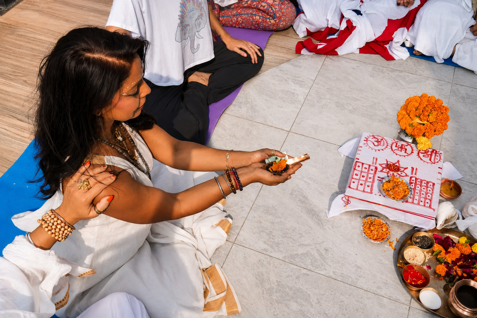 A woman participating in a traditional Hindu prayer ceremony, surrounded by flowers, candles, and religious symbols, with others sitting nearby.