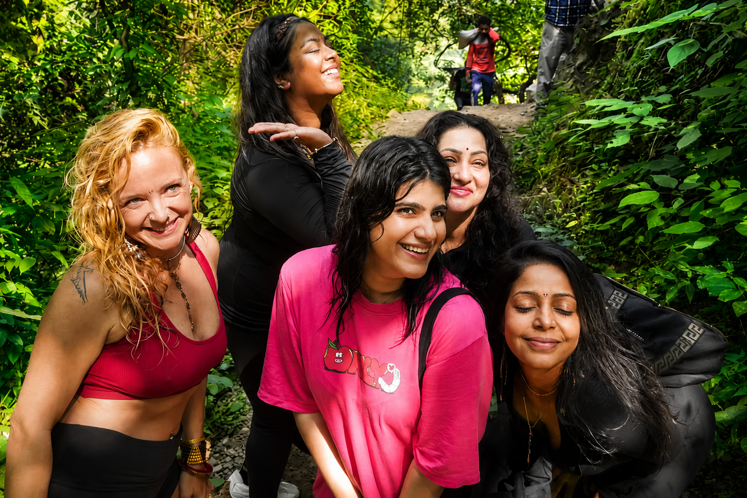 A group of six smiling women, of diverse ethnicities, enjoying a hike through a lush green forest on a sunny day.