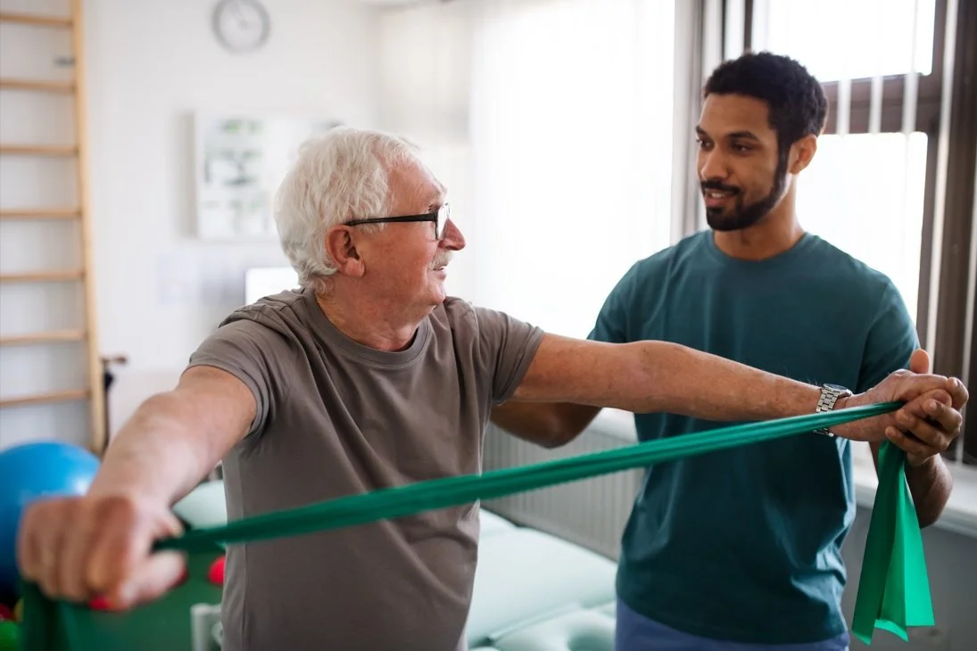 A young man helping an elderly man with physical therapy exercises using a resistance band in a therapy room.