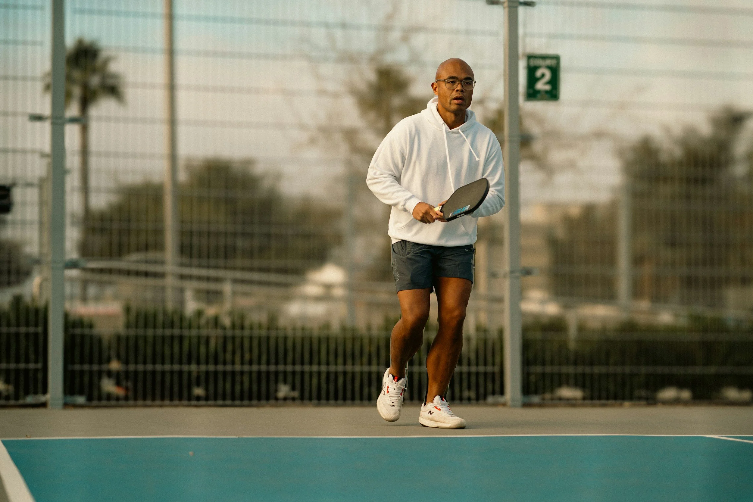A man holding a pickleball paddle on an outdoor pickleball court with a chain-link fence and trees in the background.