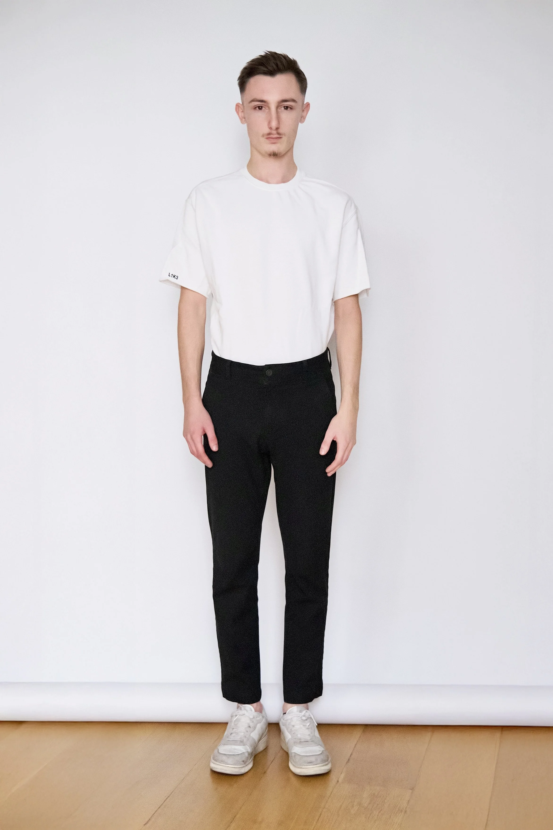 Young man standing against a white background, wearing a white t-shirt, black pants, and white sneakers.