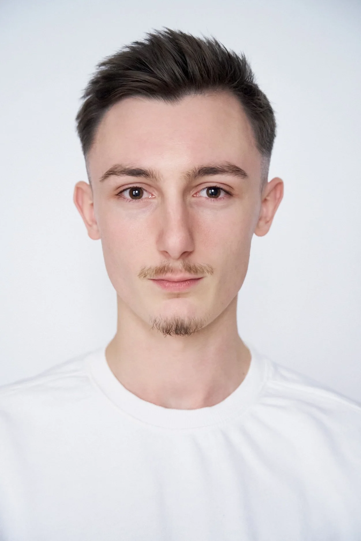 Close-up portrait of a young man with short, styled hair, light facial hair, and wearing a white shirt, against a plain white background.