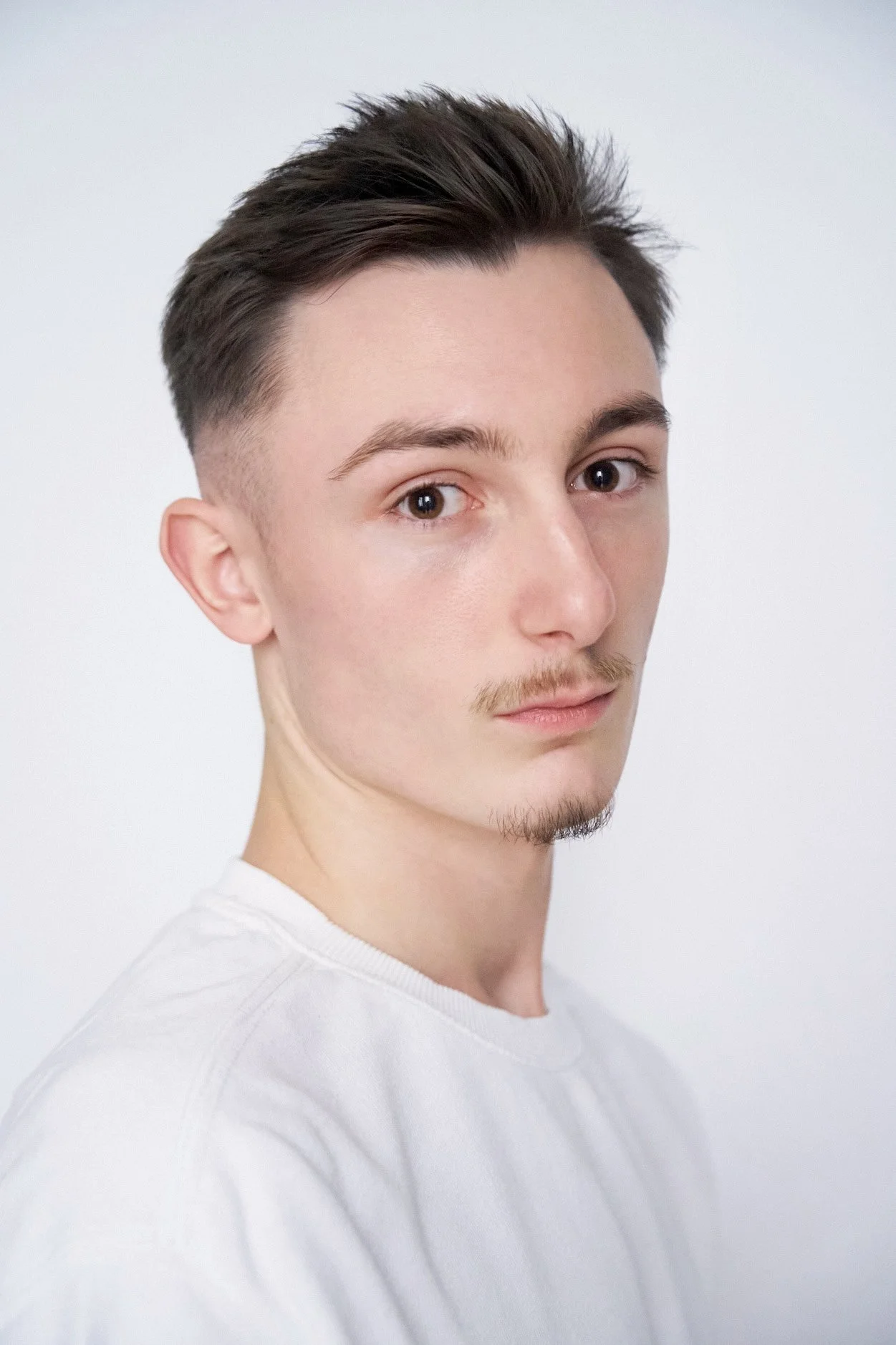 Close-up portrait of a young man with brown hair, hazel eyes, a light mustache, and a small goatee, wearing a white shirt, against a plain white background.