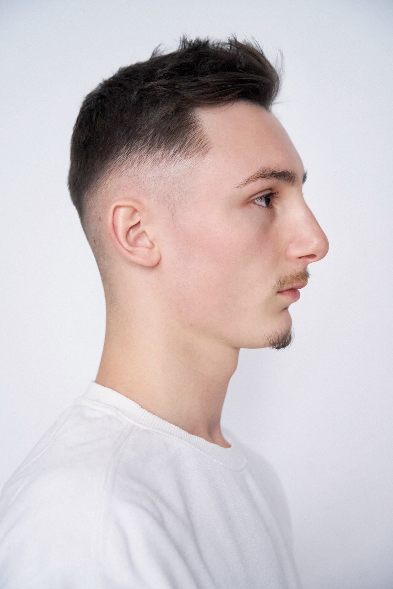 Profile of a young man with a clean fade haircut, light facial hair, and wearing a white T-shirt, against a plain white background.