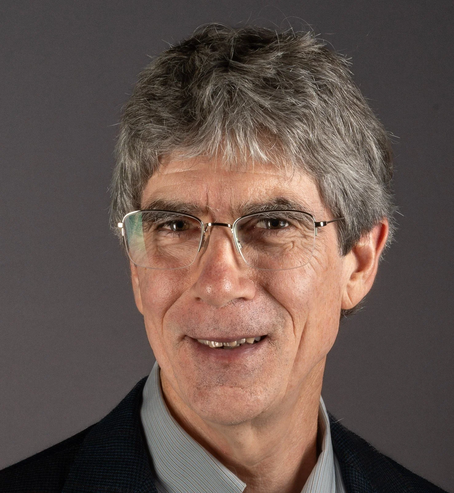 Close-up portrait of a middle-aged man with gray hair, glasses, wearing a dark blazer and a striped shirt, against a gray background.